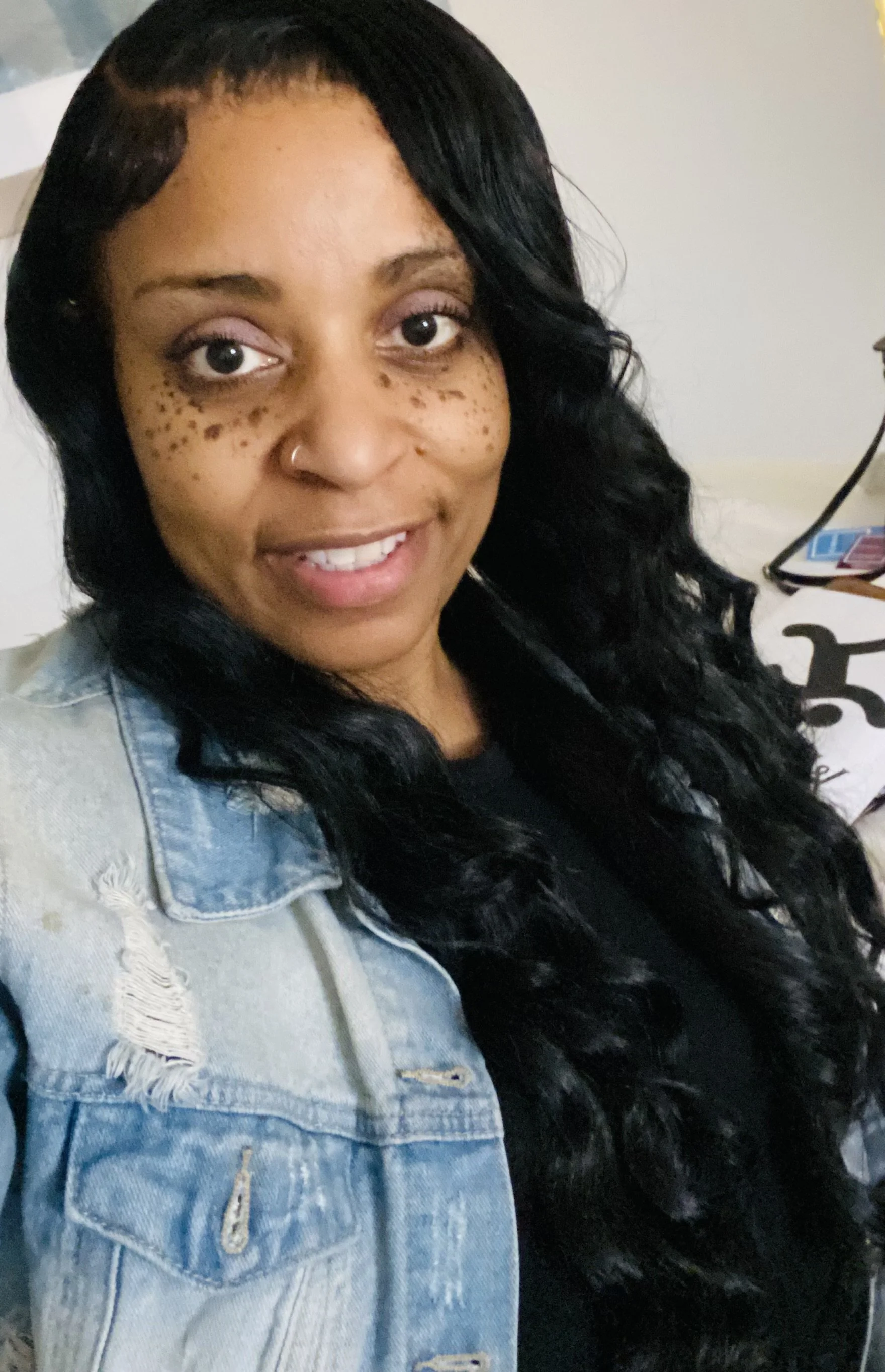 A woman with long black curly hair, freckles, a nose piercing, and wearing a denim jacket smiling at the camera.