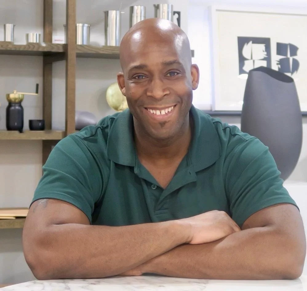 A smiling man with a bald head and a beard sitting at a table in a modern office or living space, with shelves and decorative items in the background.