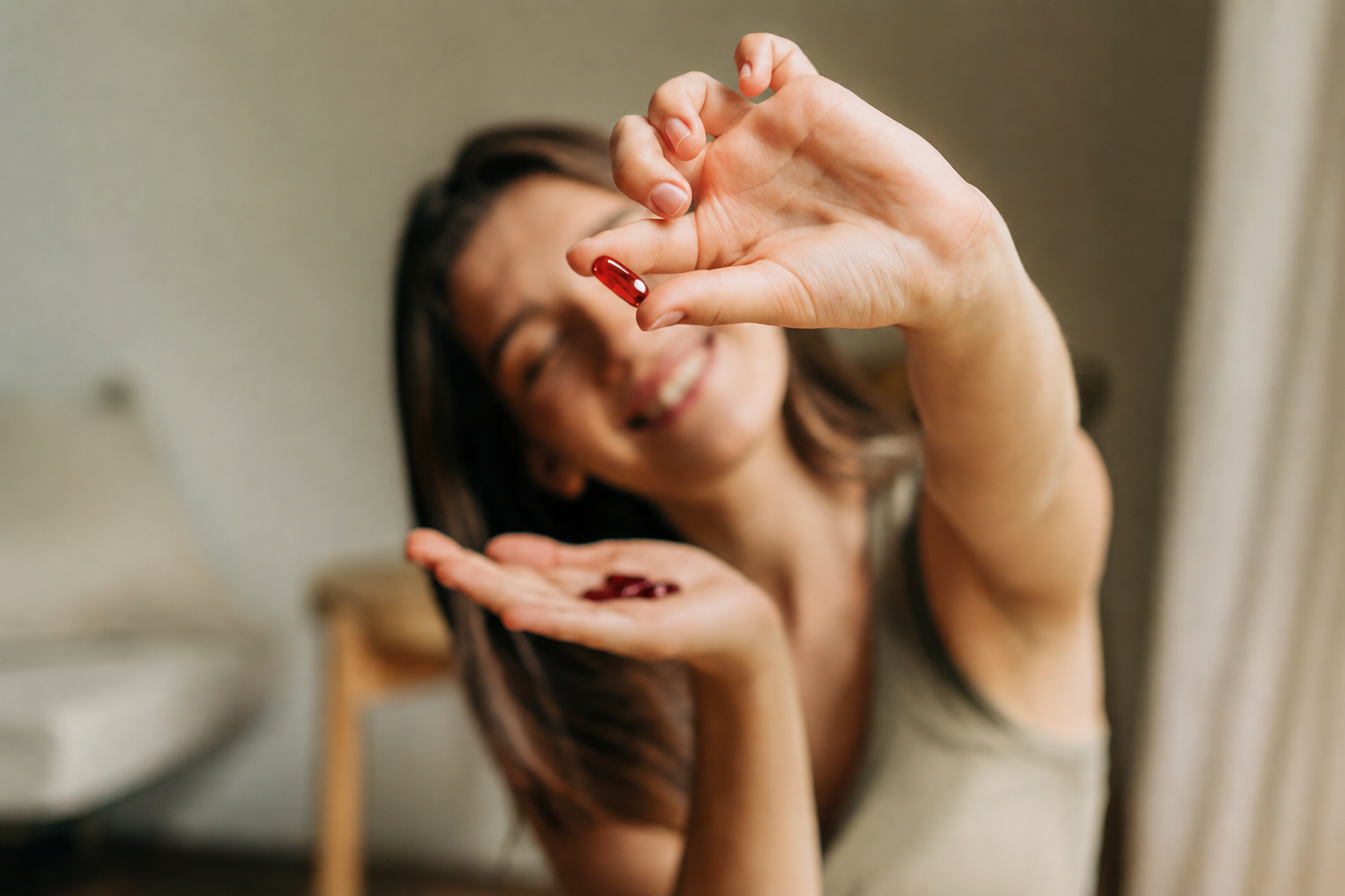 Smiling woman holding a red capsule pill in her hand, with another pill in her open palm, blurred background.