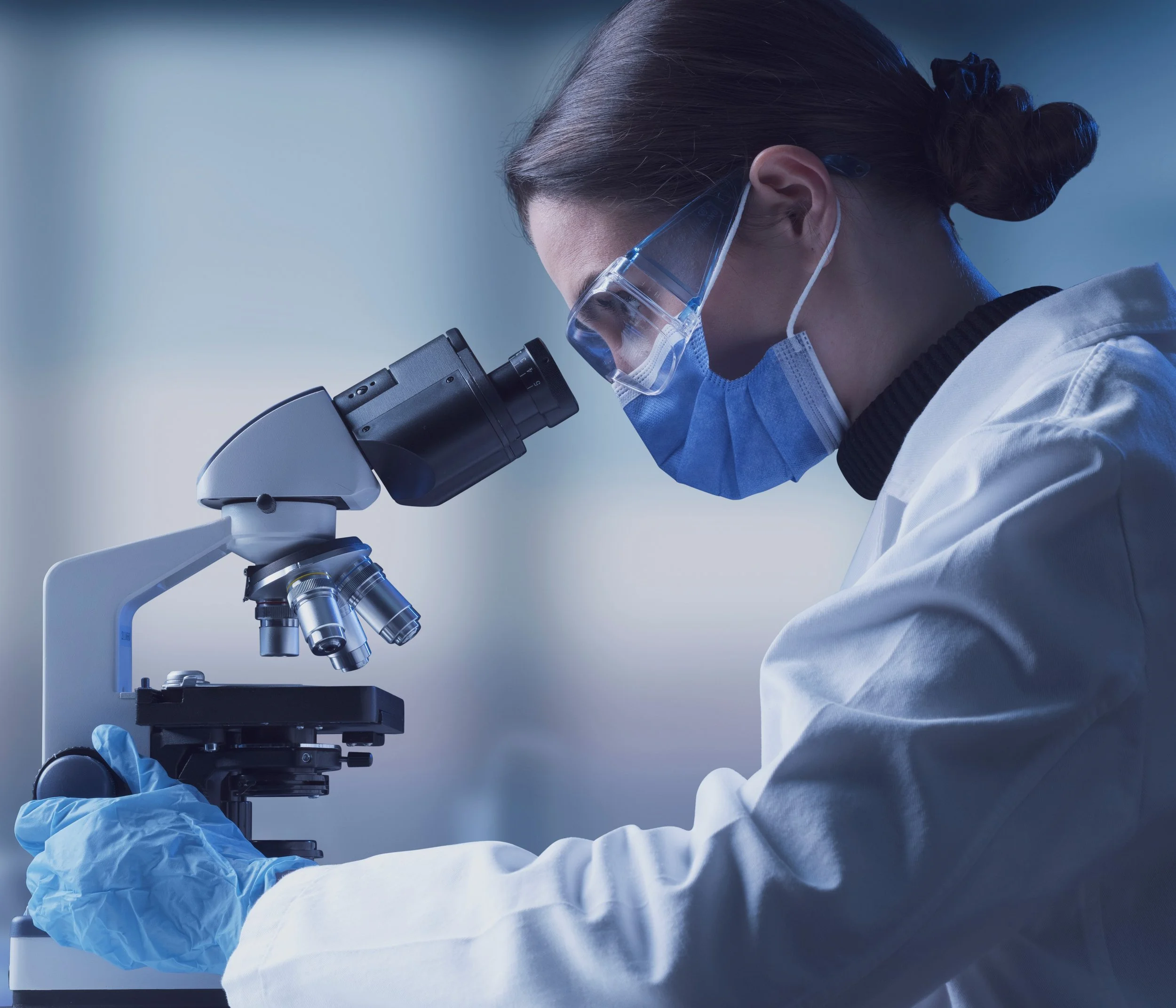 A scientist or researcher wearing safety goggles, a face mask, gloves, and a lab coat looking through a microscope in a laboratory.