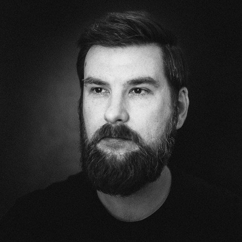 Portrait en noir et blanc d'un homme avec une barbe et des cheveux courts, regardant légèrement vers la droite, fond sombre.
