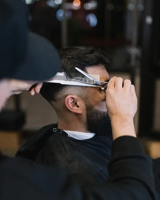 Un homme avec une barbe qui reçoit une coupe de cheveux dans un salon de coiffure, le coiffeur utilise des ciseaux et une règle pour tailler sa coupe.