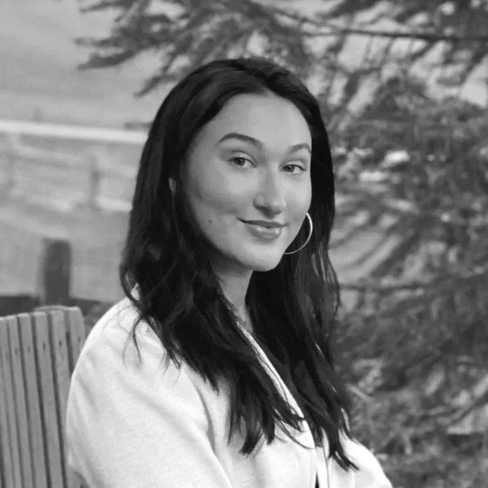 This is a black and white photo of Hannah Lukhard. She has black hair and is seated on a bench in front of a tree. She is wearing a white top.
