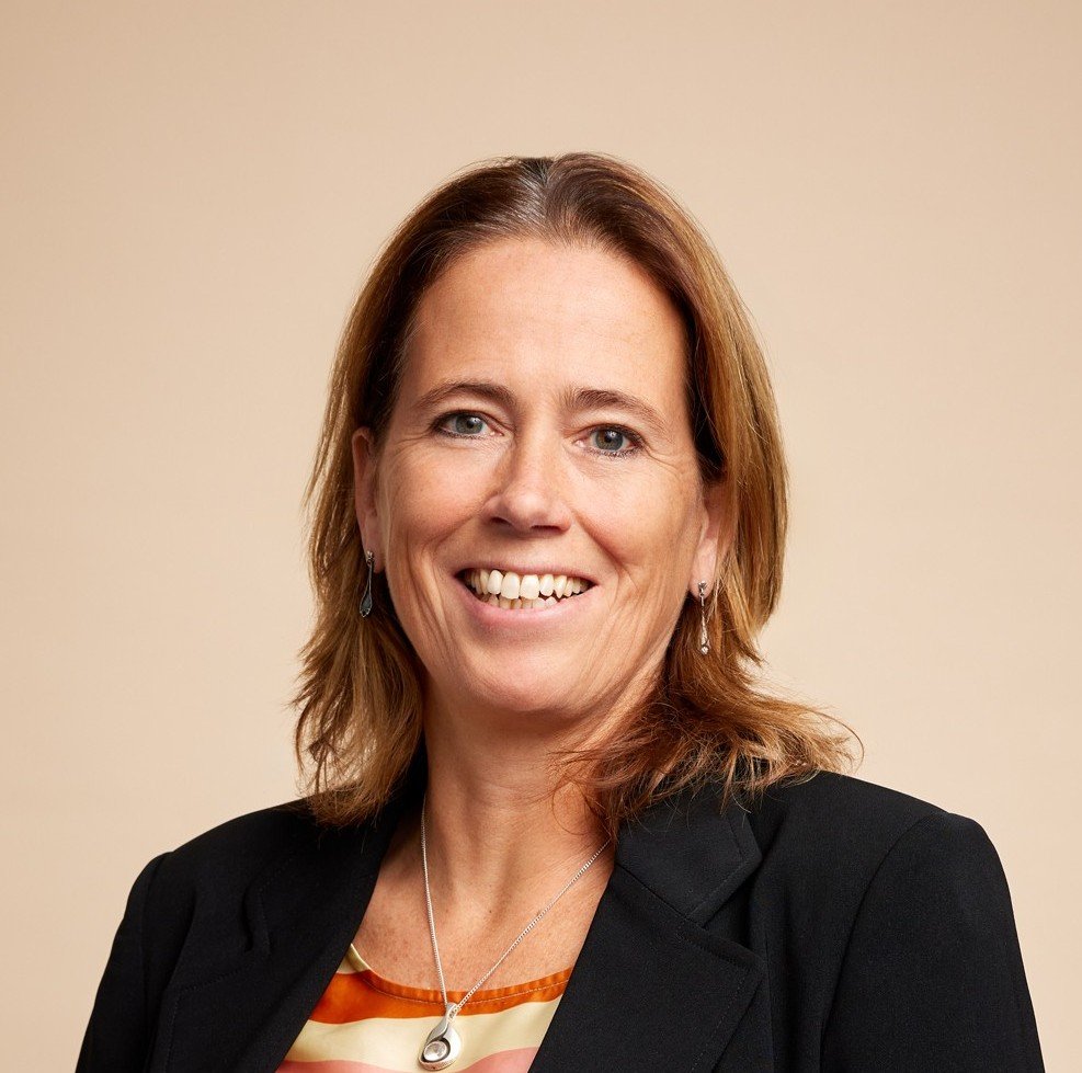 A smiling woman with shoulder-length brown hair, wearing a black blazer, colorful top, earrings, and a necklace, against a beige background.