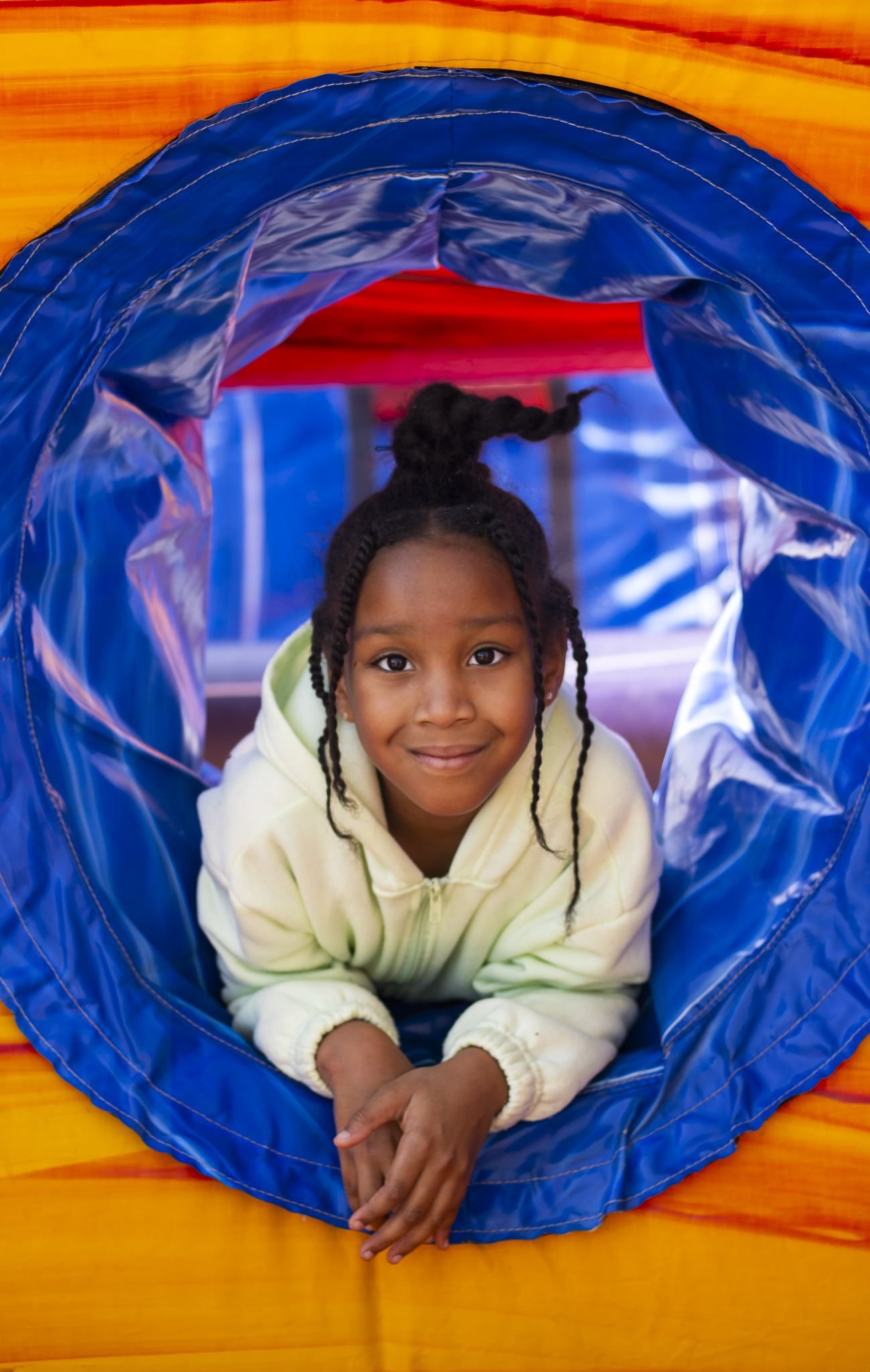 A young girl with braided hair smiling inside a blue and orange play tunnel.