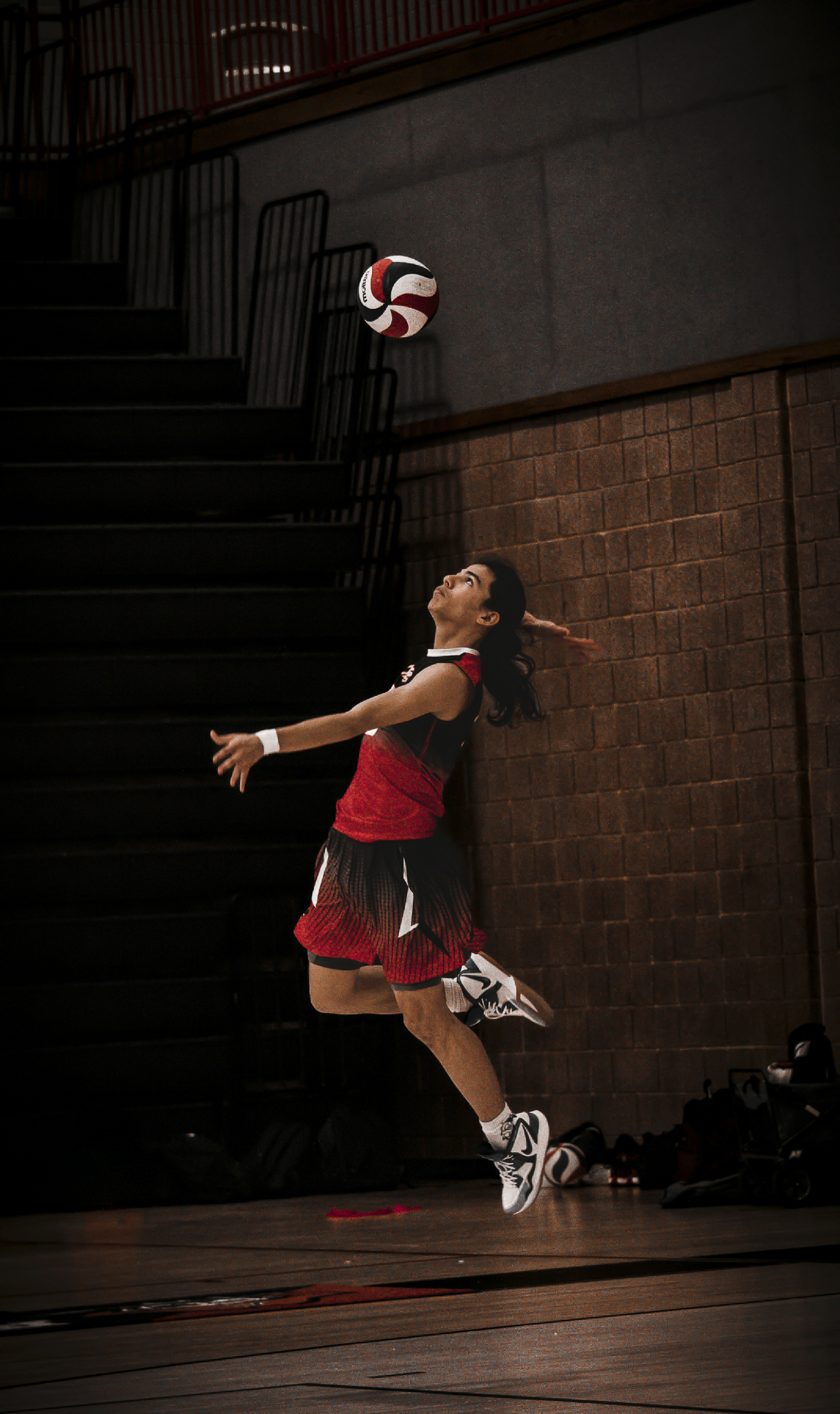 A female volleyball player in red and black uniform jumping to hit a volleyball indoors with wooden flooring and brick wall background.