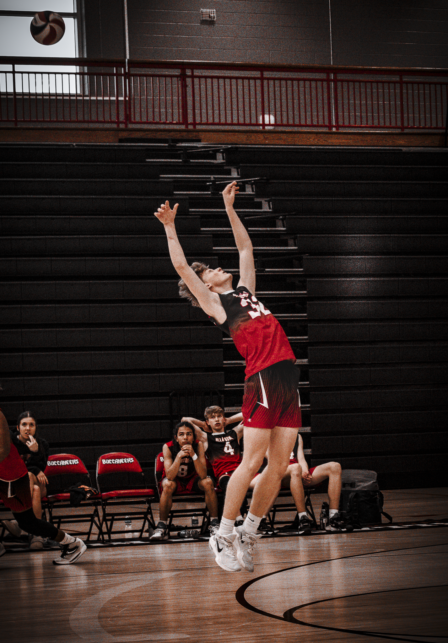 A volleyball player in a red and black uniform jumps to hit the ball during a game, with teammates sitting on a bench in the background.