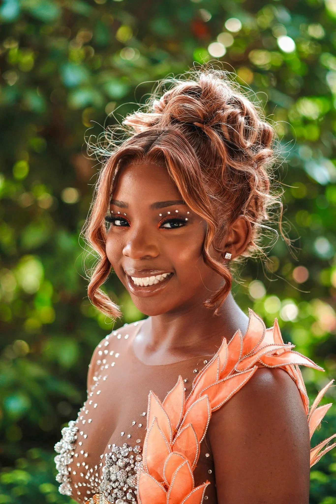 Young woman with styled hair, adorned with rhinestones and floral embellishments on her dress, smiling outdoors against a blurred green background.