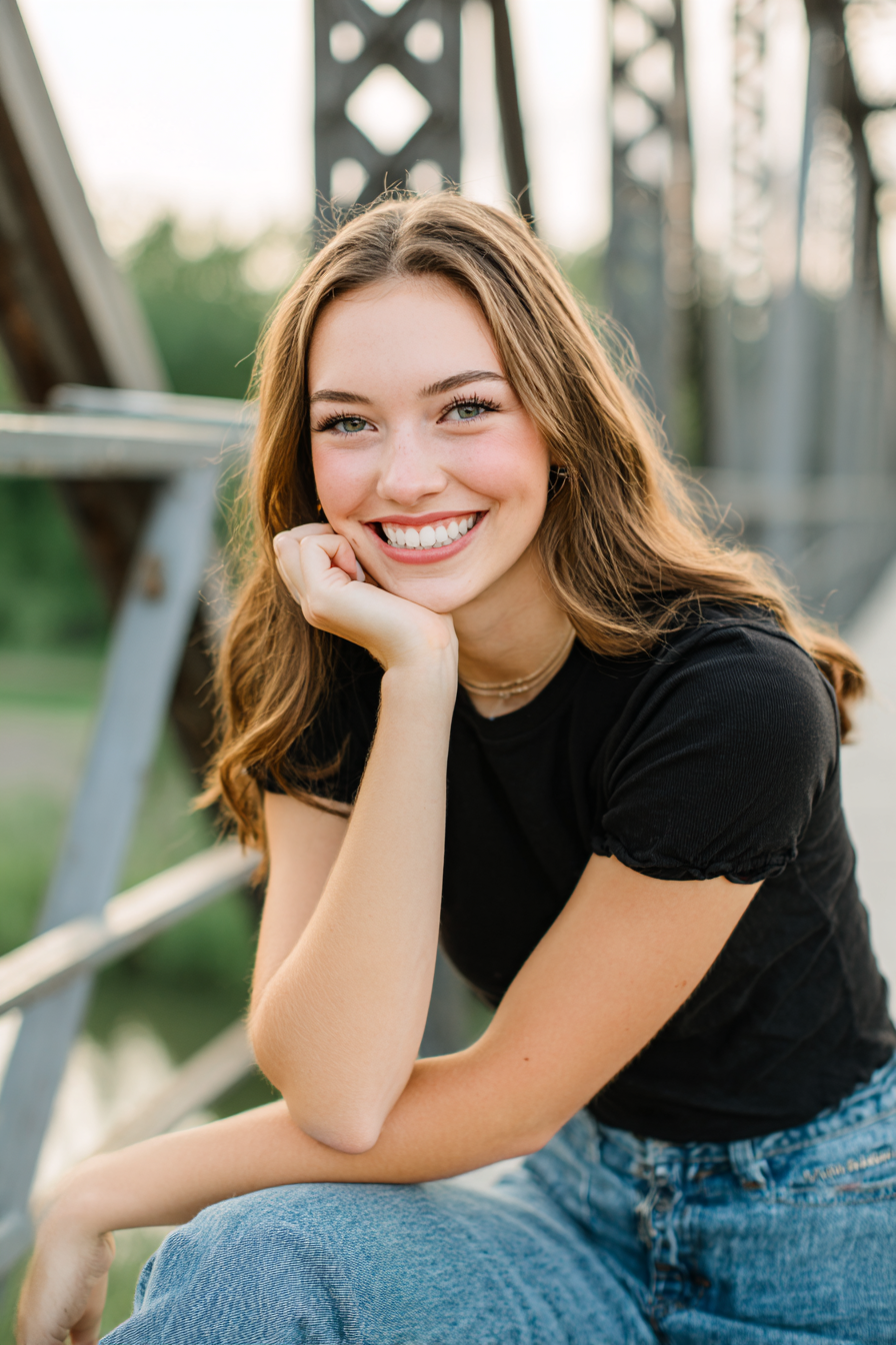 Close-up of a smiling young woman with light brown hair, wearing a black t-shirt, outdoors on a bridge at sunset.