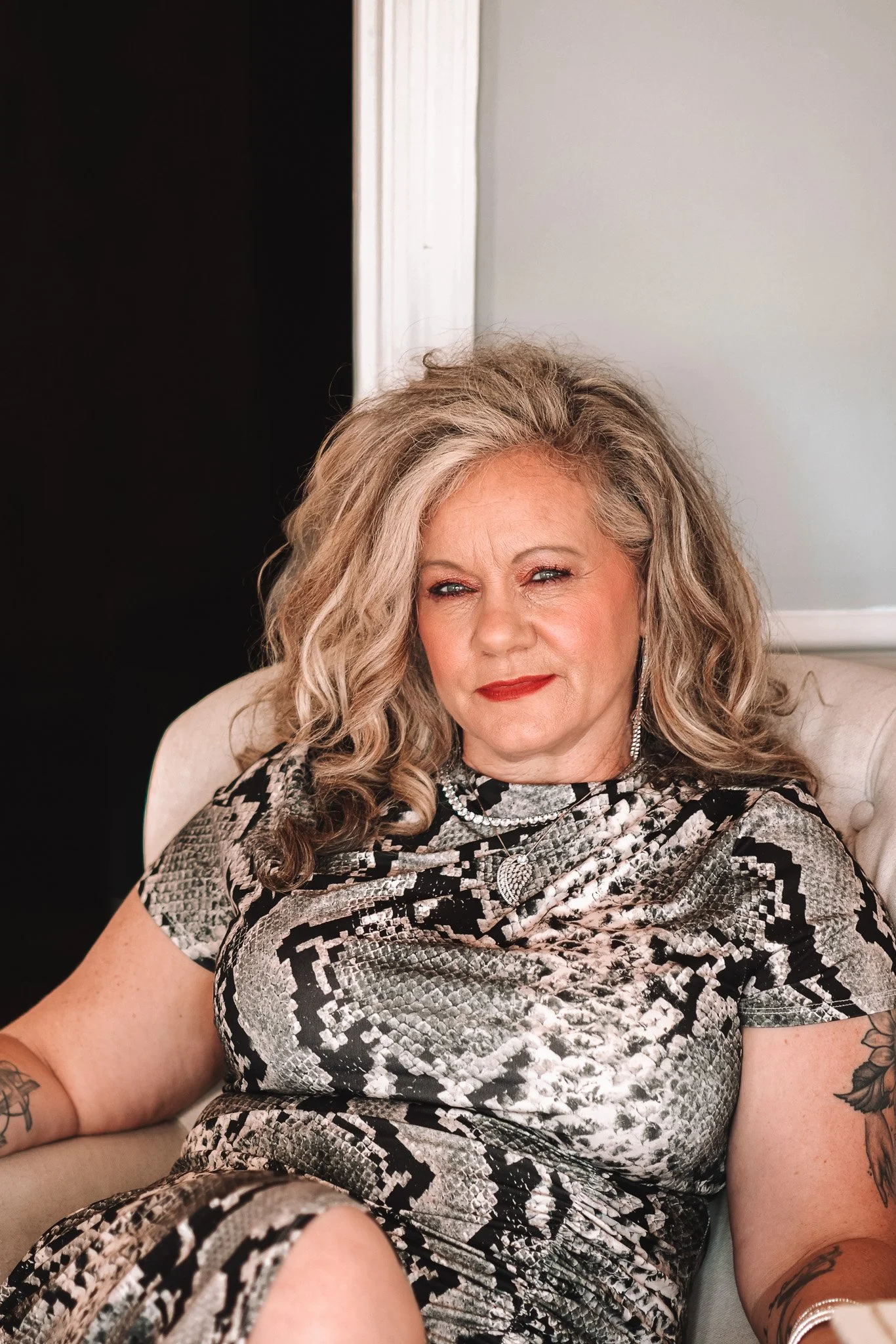 A mature woman with curly gray hair sits on a beige couch, wearing a snake print dress and jewelry, with tattoos on her arms, looking confidently at the camera.