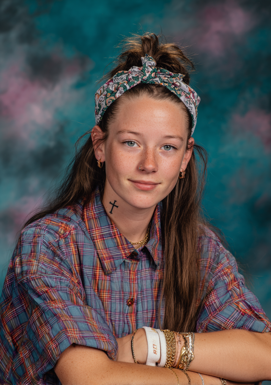 A young woman with long brown hair wearing a plaid shirt and a patterned headband, smiling slightly, with a cross tattoo on her neck, multiple earrings, and layered bracelets on her wrist, against a colorful, cloud-like background.