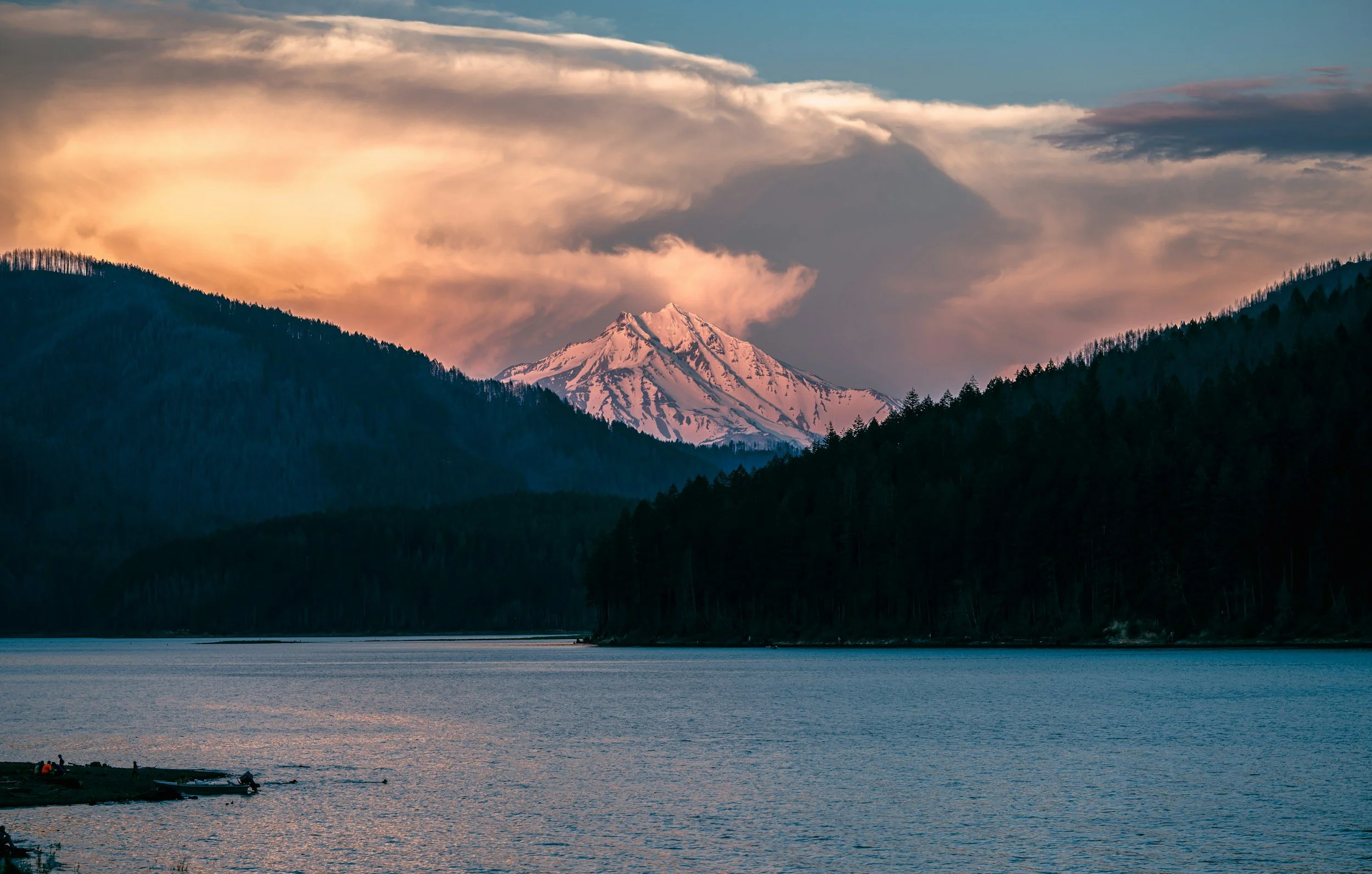 Mount Rainier snow-capped mountain in the distance with detroit lake and forested hills in the foreground during sunset.