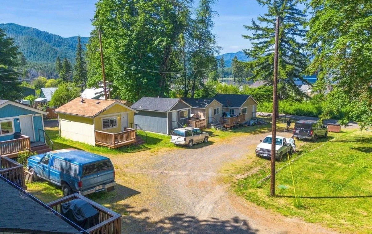 A small residential area with a gravel parking lot, several small houses, parked vehicles, and large green trees. Mountains and a blue sky are seen in the background.