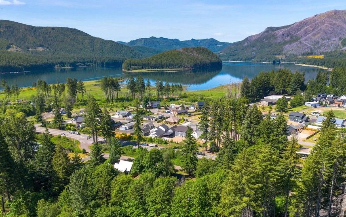 A scenic aerial view of a small residential area surrounded by lush green trees, with a large lake reflecting the blue sky and mountains in the background.