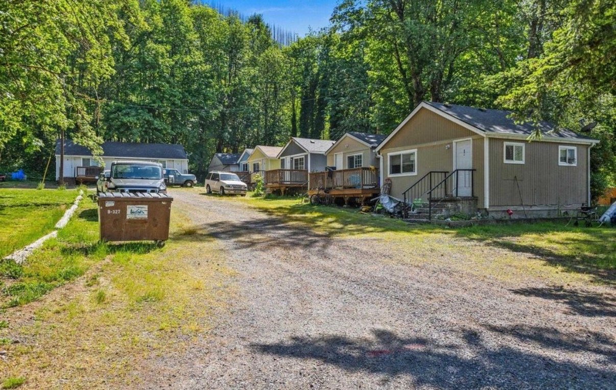 A row of small houses in a wooded area on a sunny day, with a gravel driveway in front and a dumpster on the left side.