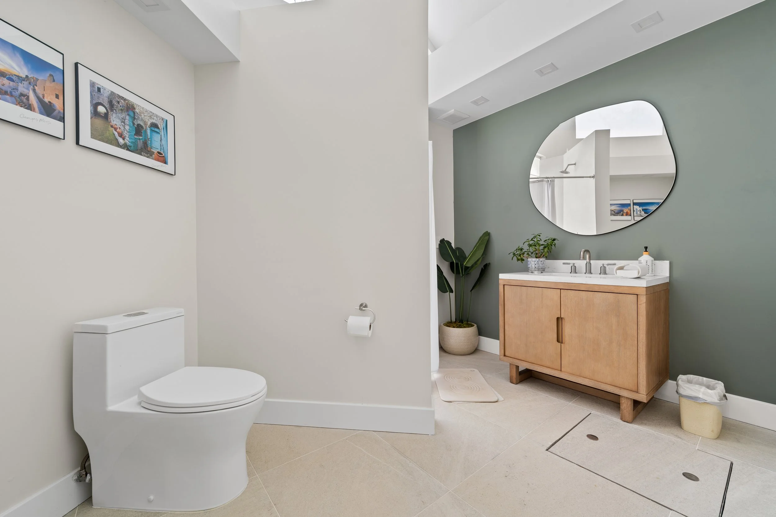 Modern bathroom with white toilet, light-colored tiled floor, white walls with framed artwork, and a vanity with a wooden cabinet, white countertop, and round mirror. There are plants, a soap dispenser, and a trash can visible.