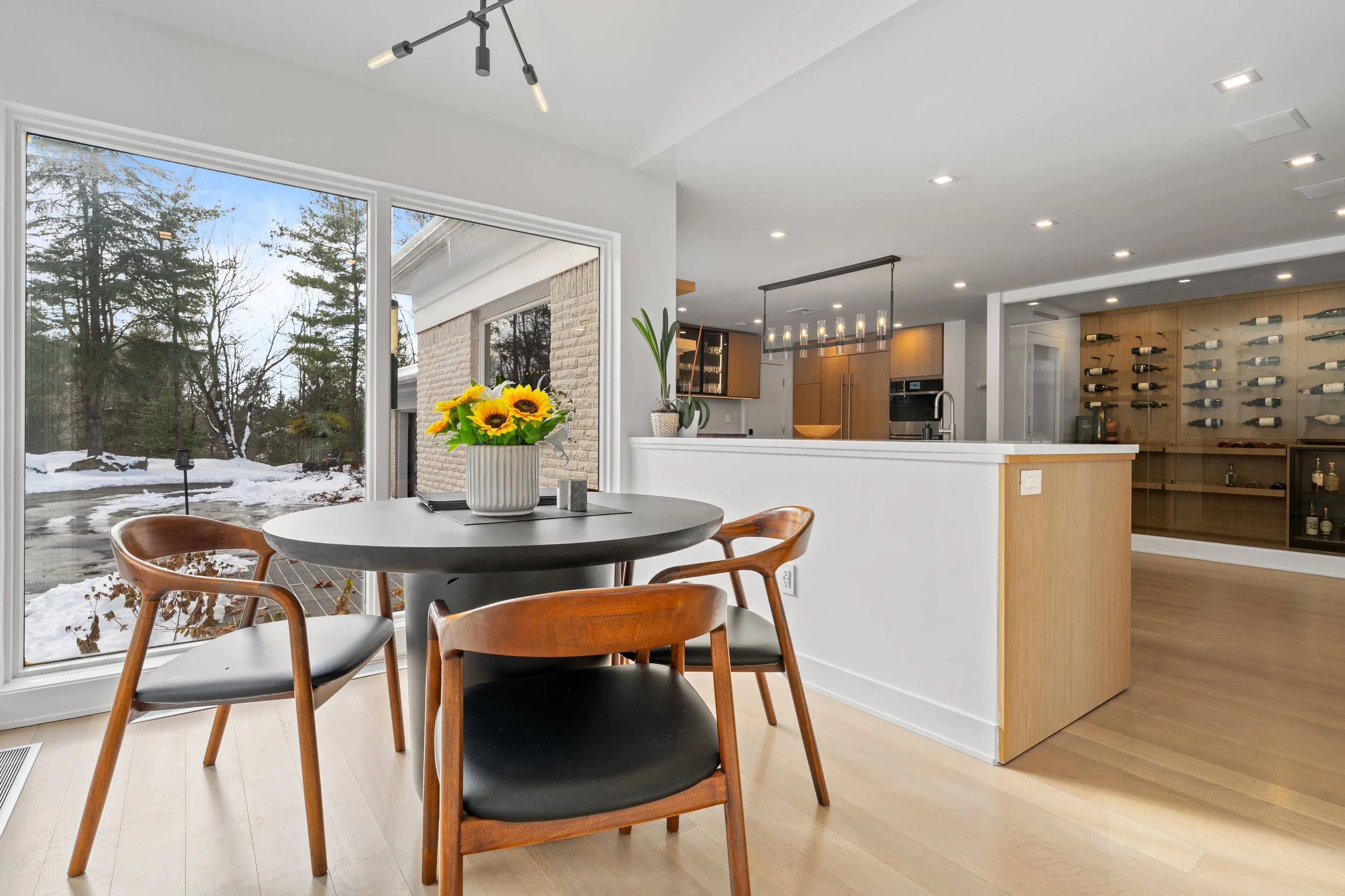 A modern dining area with a round table, four wooden chairs with black seats, and a flower arrangement with sunflowers on the table. Large windows showing a snowy outdoor scene, adjacent to a kitchen with wooden cabinets and a wine rack.