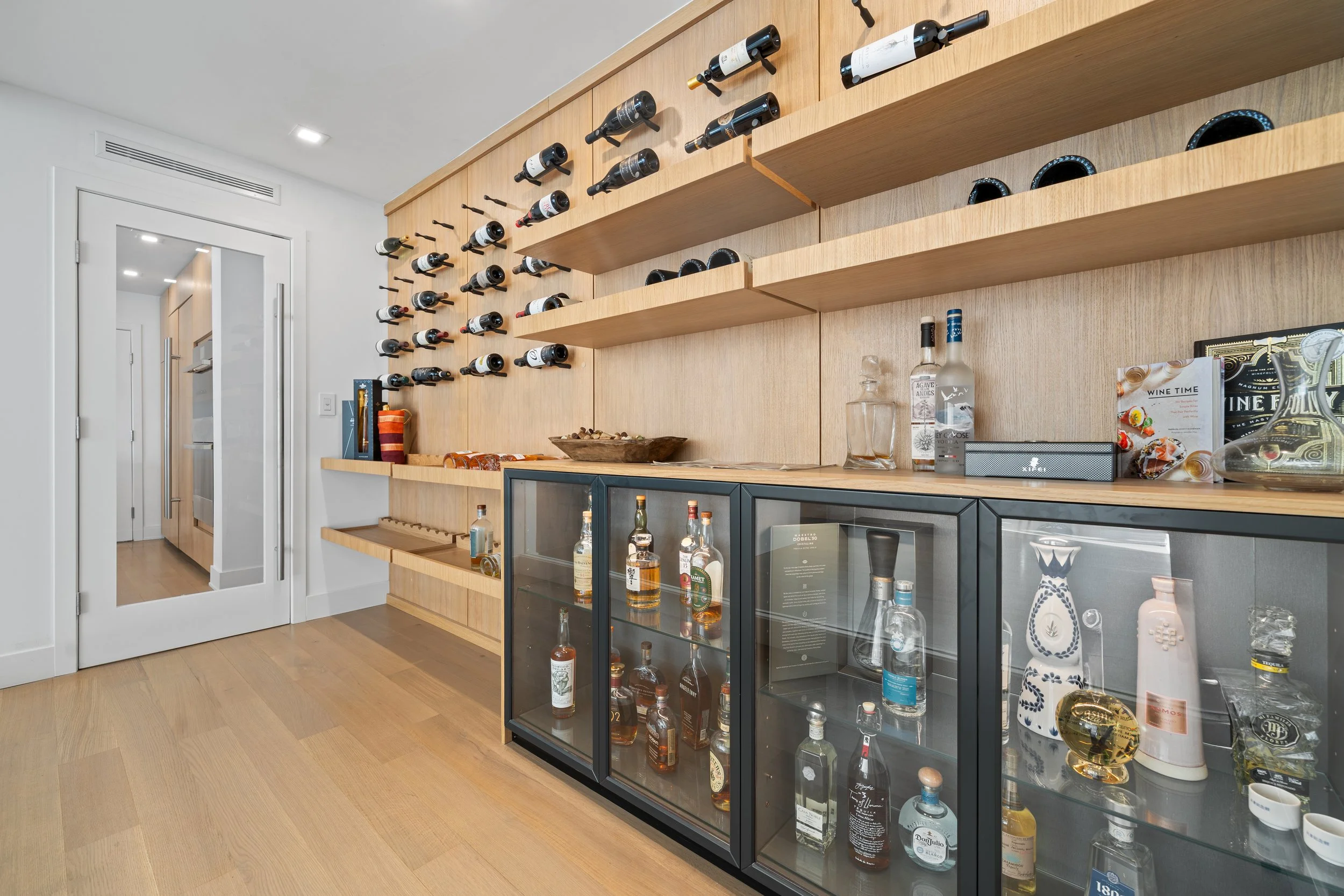Home bar area with wooden shelves holding wine bottles, a glass-fronted cabinet with liquor bottles, and a glass pitcher, in a modern room with wooden flooring and a door with a full-length mirror.