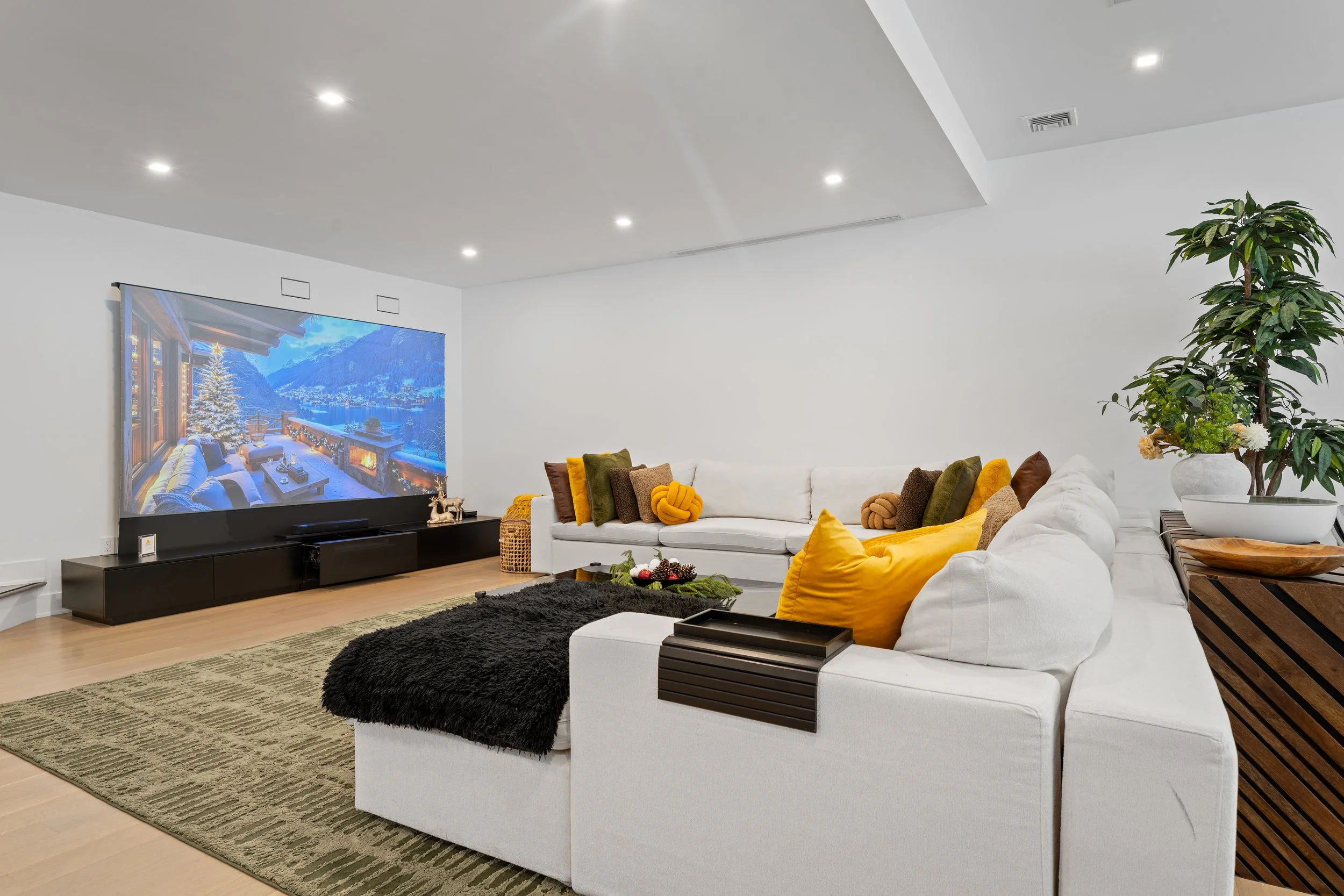 Living room with a large flat-screen TV showing a winter snowy scene, white sectional sofa with yellow, brown, and green pillows, a black rug, wooden side table with a large potted plant, and a green rug on light hardwood floors.