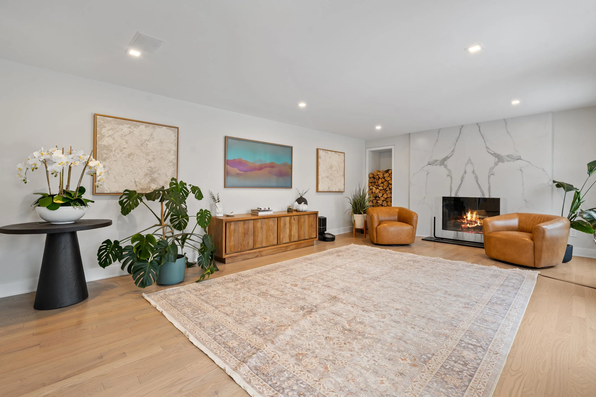 Modern living room with a fireplace, two brown leather chairs, a wooden sideboard, framed art on white walls, plants, and a large area rug.