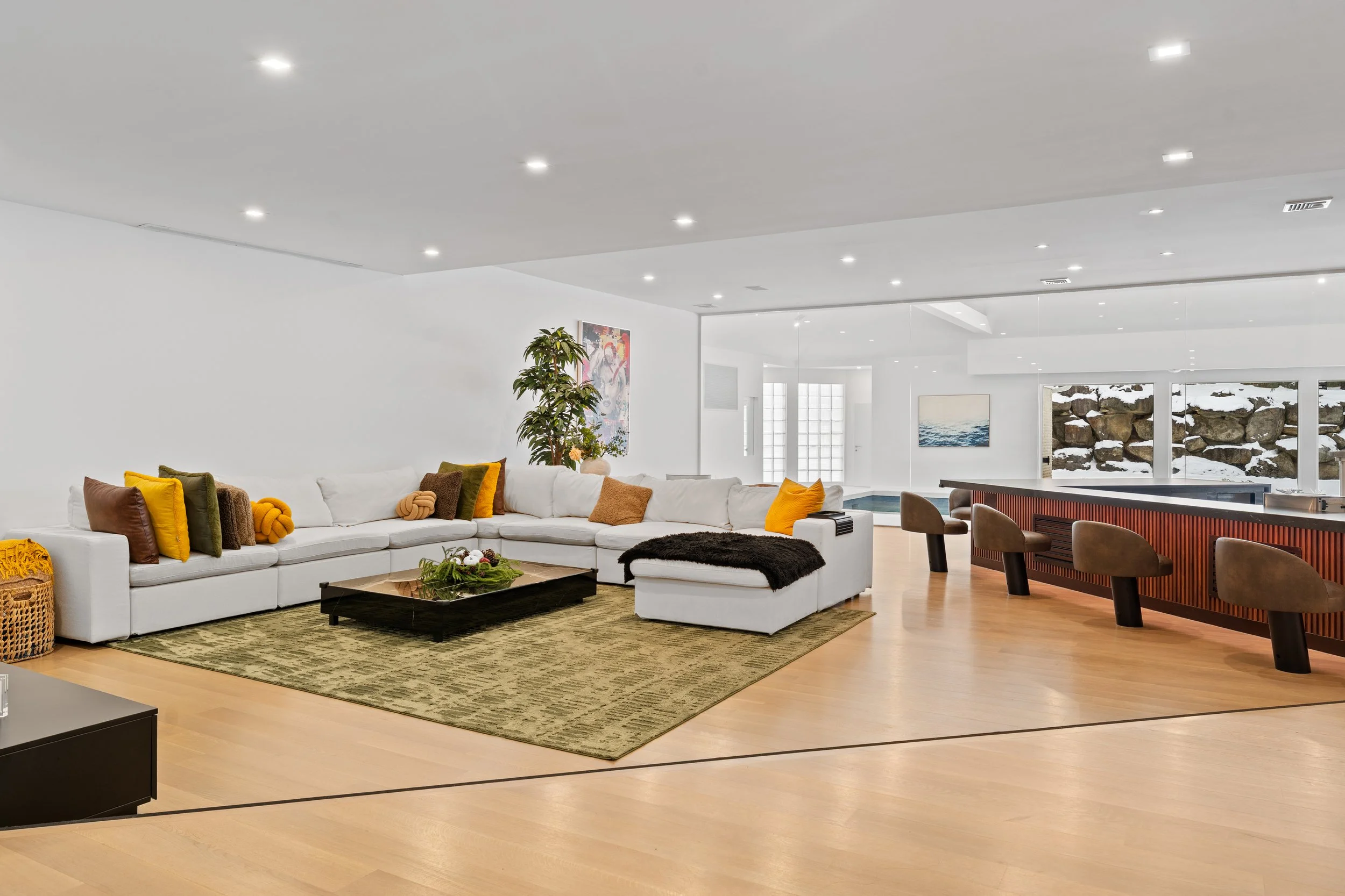 Spacious modern living room with white sectional sofa, colorful pillows, green rug, coffee table, indoor plant, art on the wall, and a bar with bar stools. Large windows show a snowy outdoor scene.