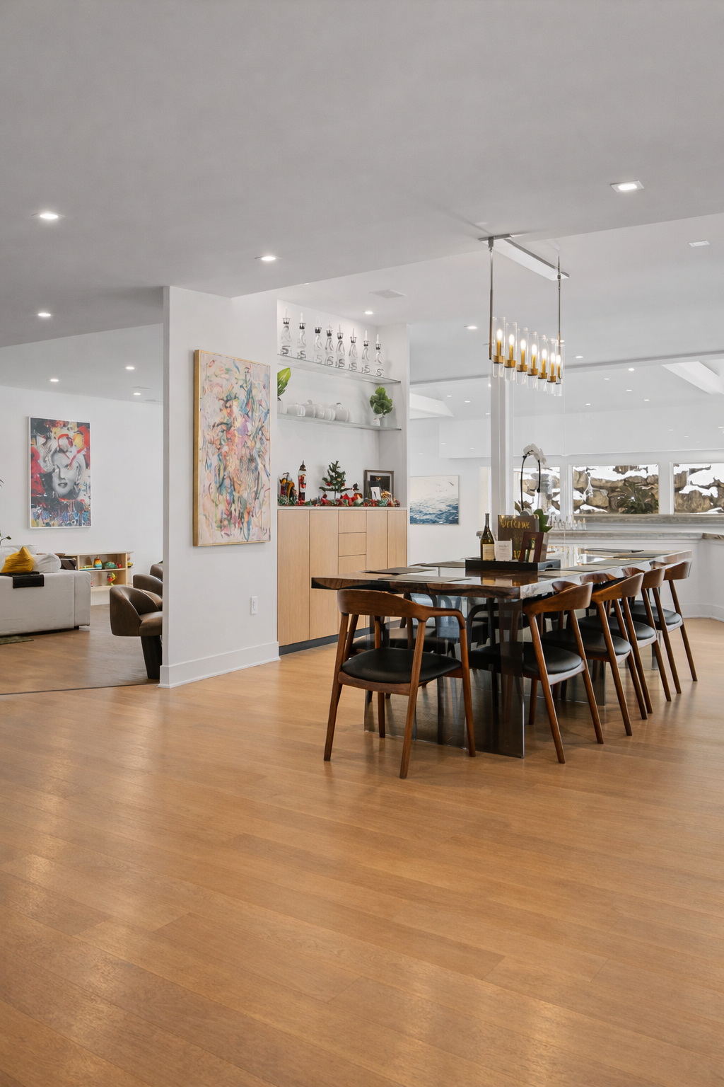 Modern dining room with wooden table and chairs, white walls decorated with colorful artwork, and a sideboard with small festive decorations, bottles, and pictures. Bright natural light from windows.
