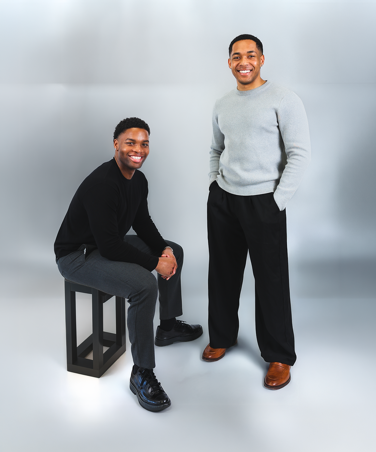 Two smiling men with dark hair and medium skin tones posing in a studio with a light gray background. One man is sitting on a black stool wearing a black shirt, gray pants, and black shoes. The other man is standing wearing a light gray sweater, black pants, and brown shoes.