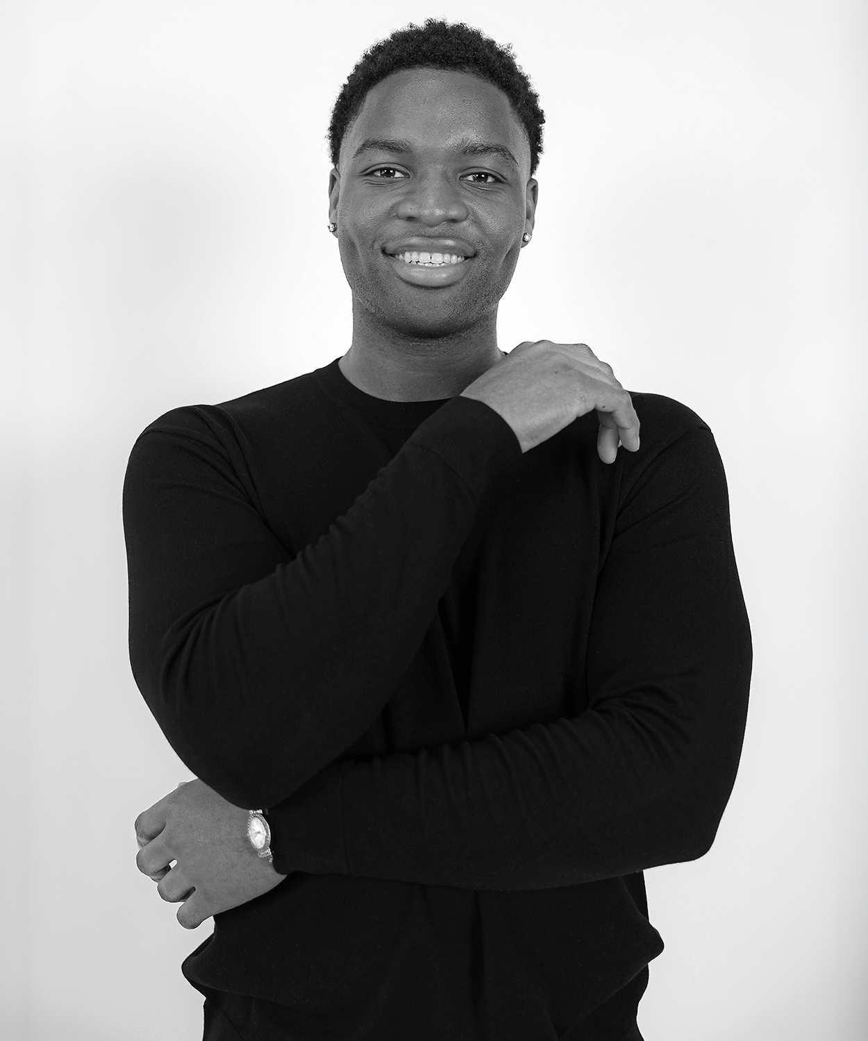 A young Black man with short curly hair, smiling, wearing a black long-sleeve shirt, with one arm crossed and the other hand resting on his shoulder, standing against a plain white background.