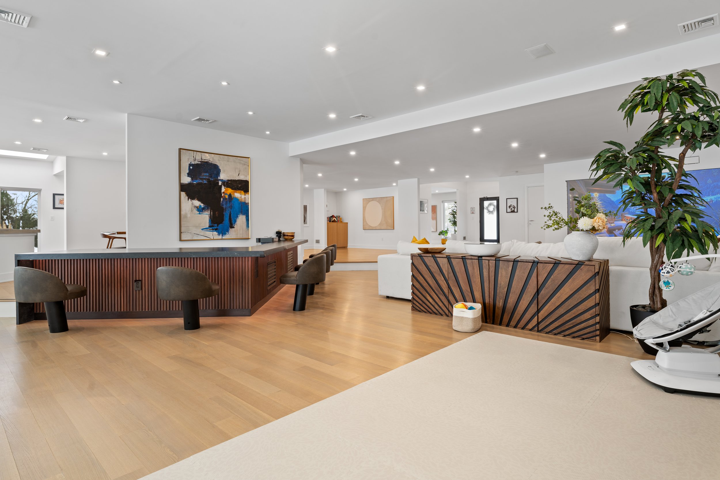 A modern, spacious living room with white walls, hardwood flooring, contemporary artwork, and minimalistic furniture, including a white sofa, wooden accents, and indoor plants.