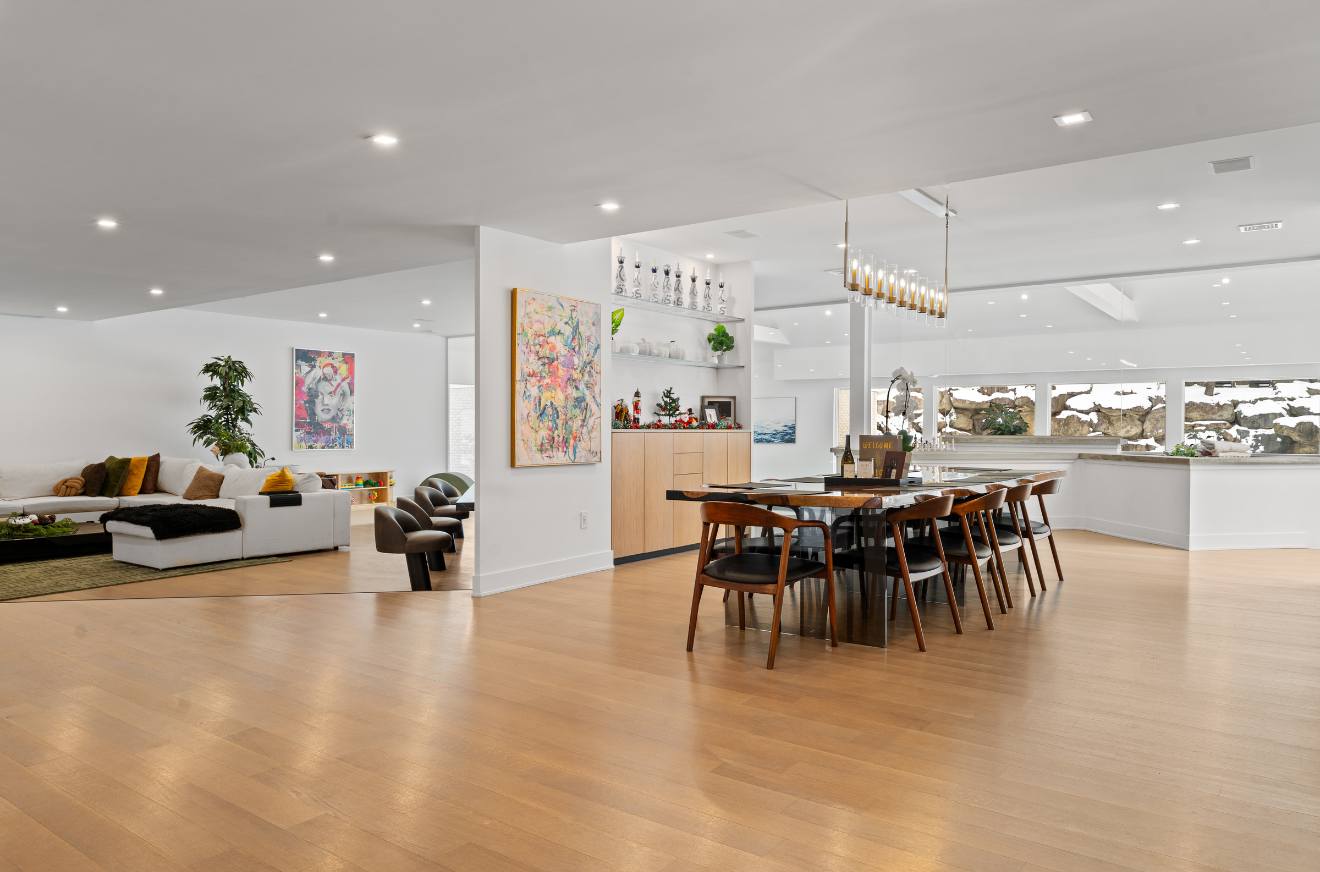 Open-concept living and dining area with hardwood floors, white walls, large window showing snow-covered rocks outside, modern furniture, abstract art, and decorative items.