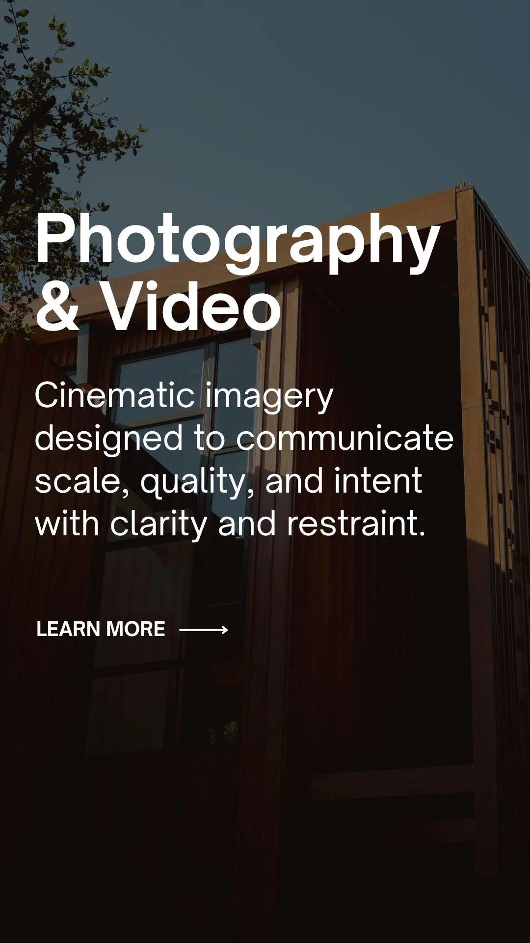 A modern house with a wooden exterior and large glass window under a clear sky, with a tree on the left.