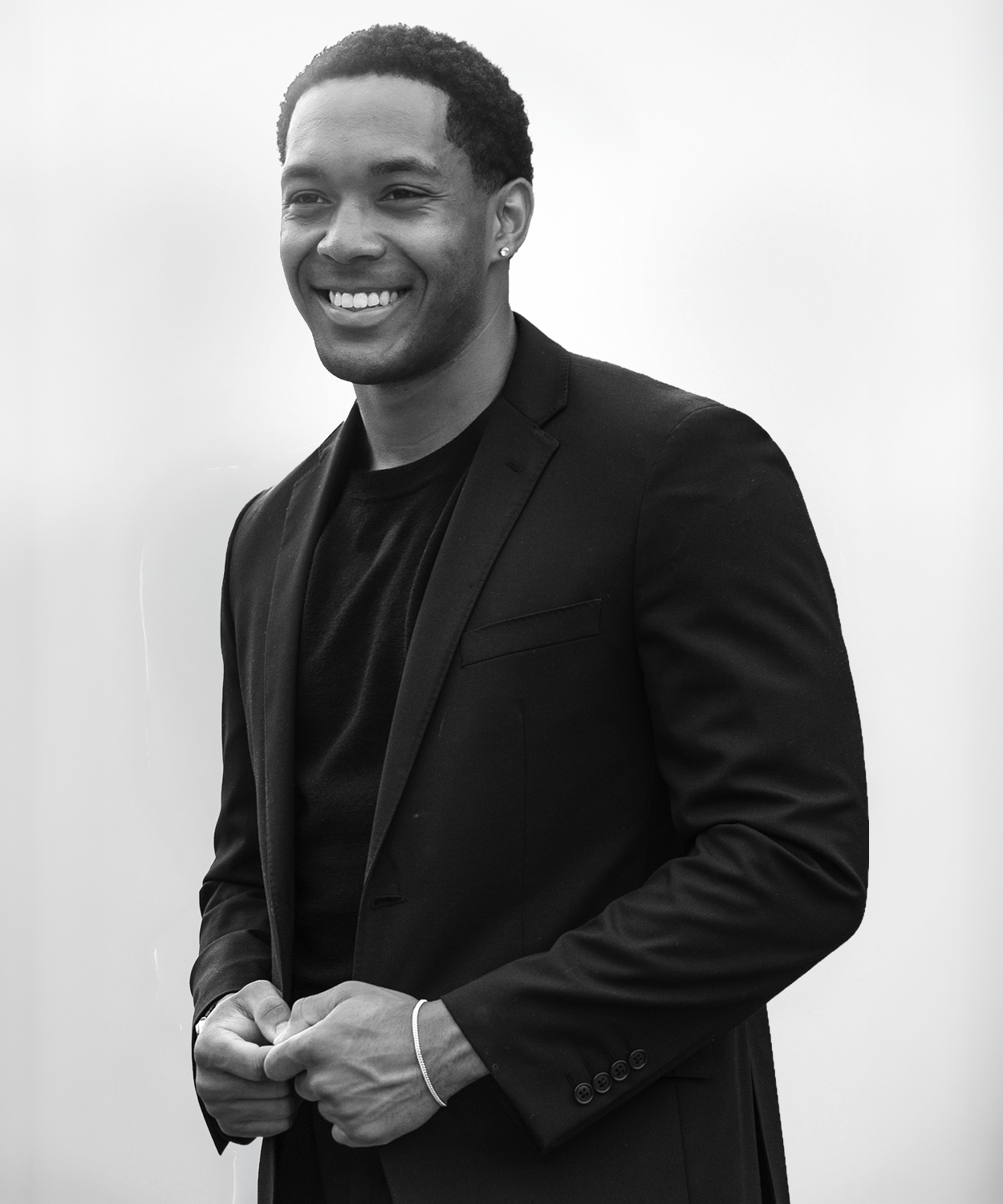 A smiling man wearing a black blazer and a black shirt, with earrings, standing against a plain background.