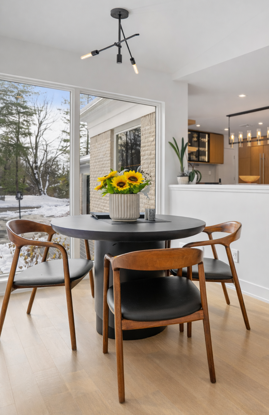 Dining area with a round black table, four wooden chairs with black cushions, a sunflower centerpiece, and large windows showing a snowy outdoor scene.
