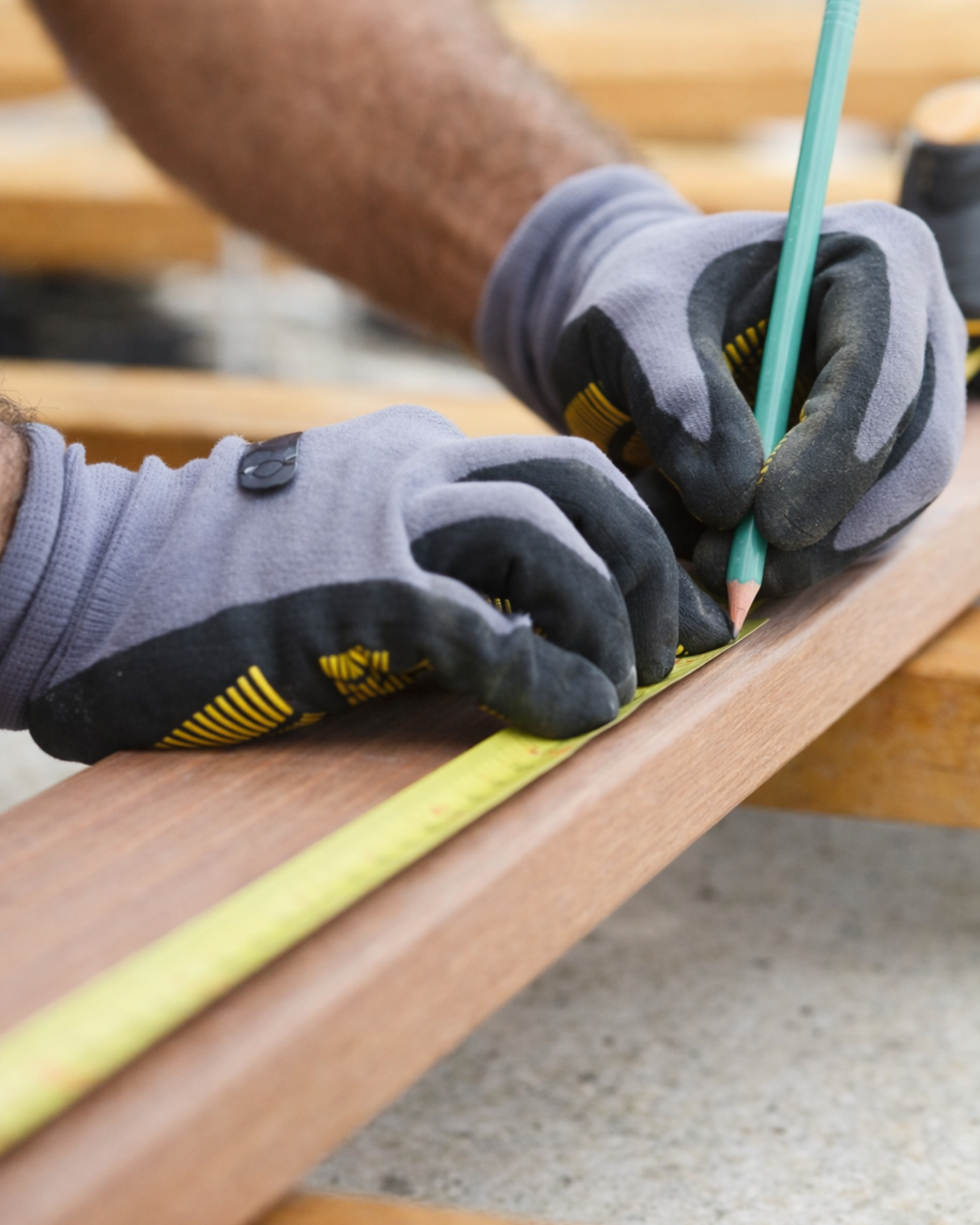A person wearing work gloves using a green pencil to measure and mark a piece of wood on a workbench.