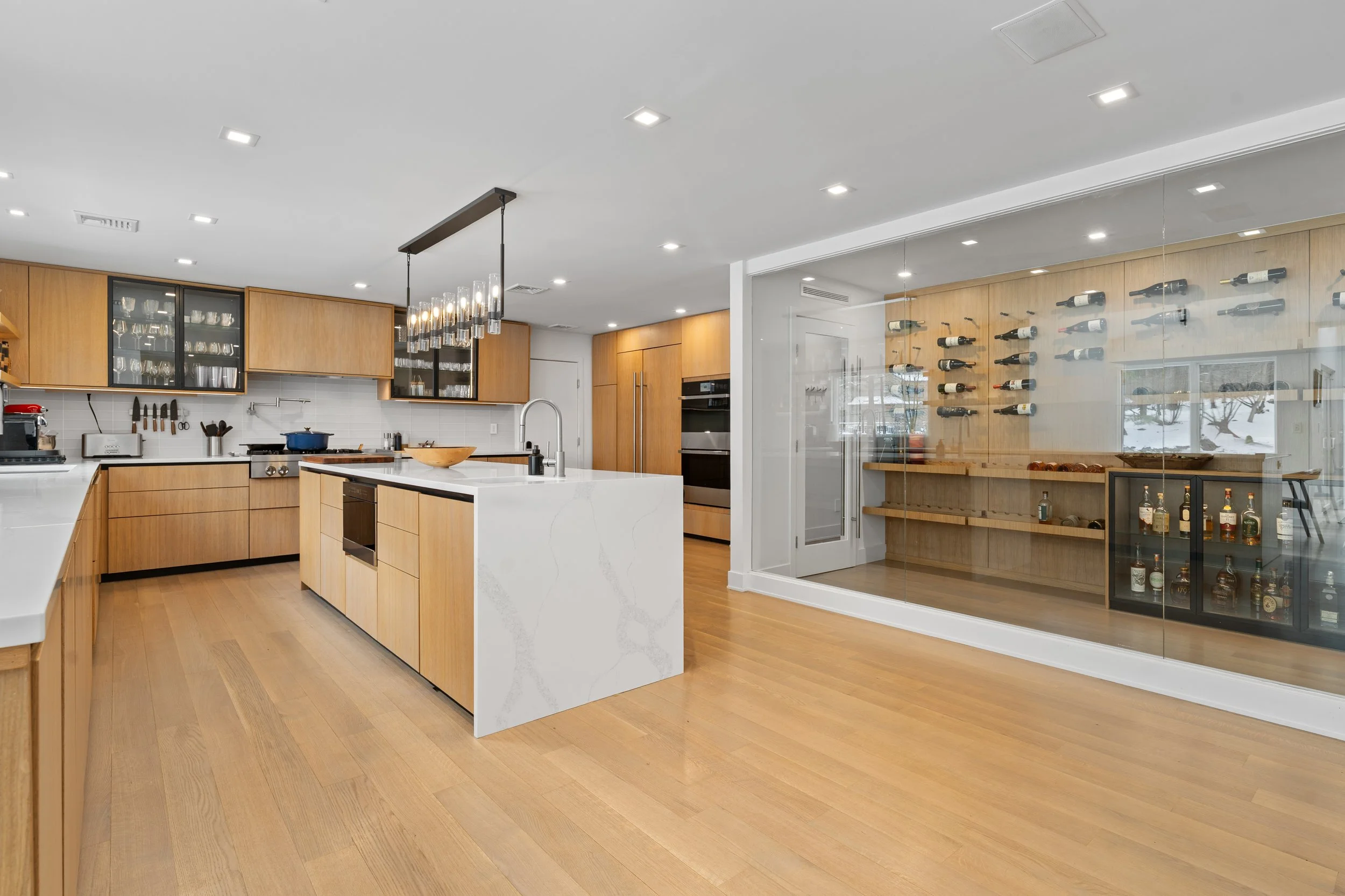 Modern kitchen with wooden cabinets, white marble island, and a glass-enclosed wine storage area.