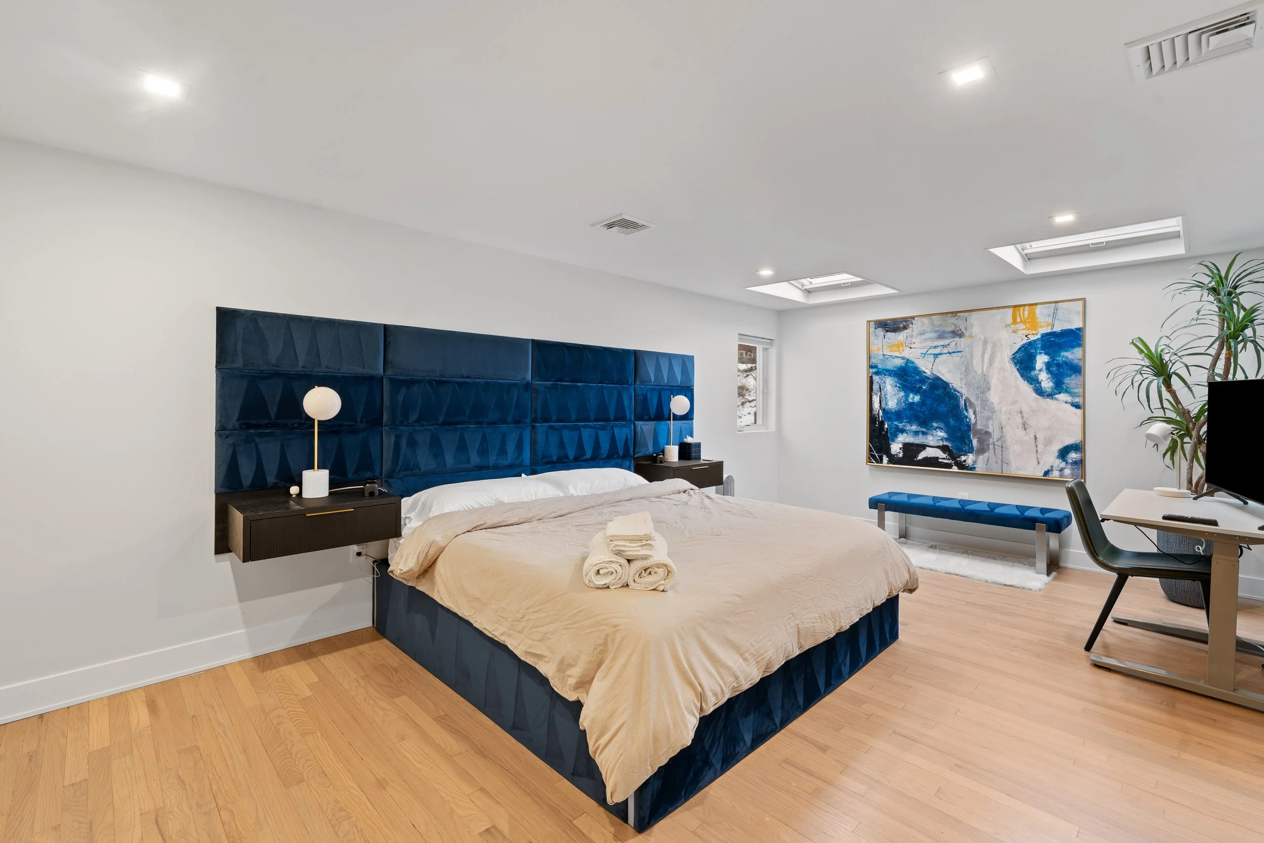 Modern bedroom with beige bedding, dark blue upholstered headboard, and wooden floors. Includes nightstands, lamps, abstract wall art, window, bench, TV, and a desk with chair.