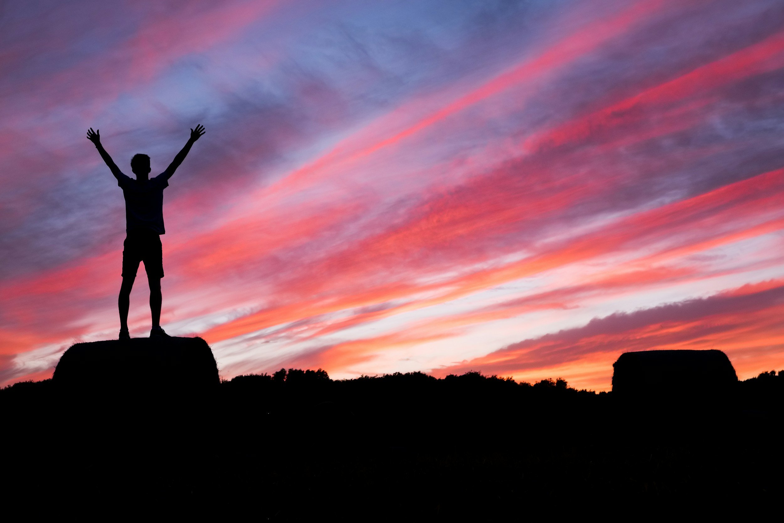 Silhouette of a person standing on a rock with arms raised at sunset, colorful sky with pink, purple, and orange clouds.