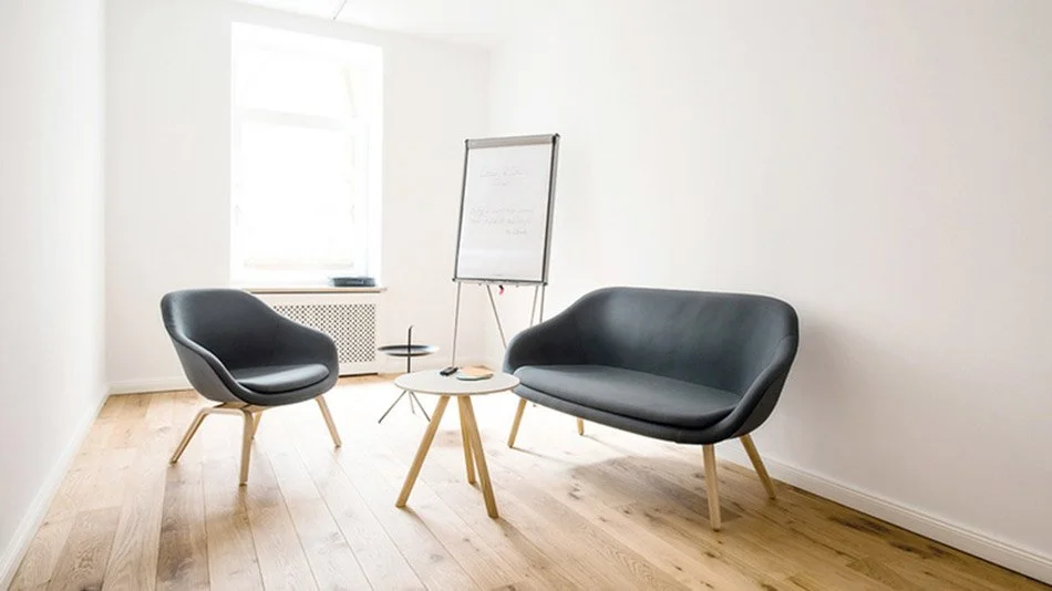 A minimal office or meeting room with two black chairs, a small round white table, a whiteboard on a stand, a window, and light wooden flooring.
