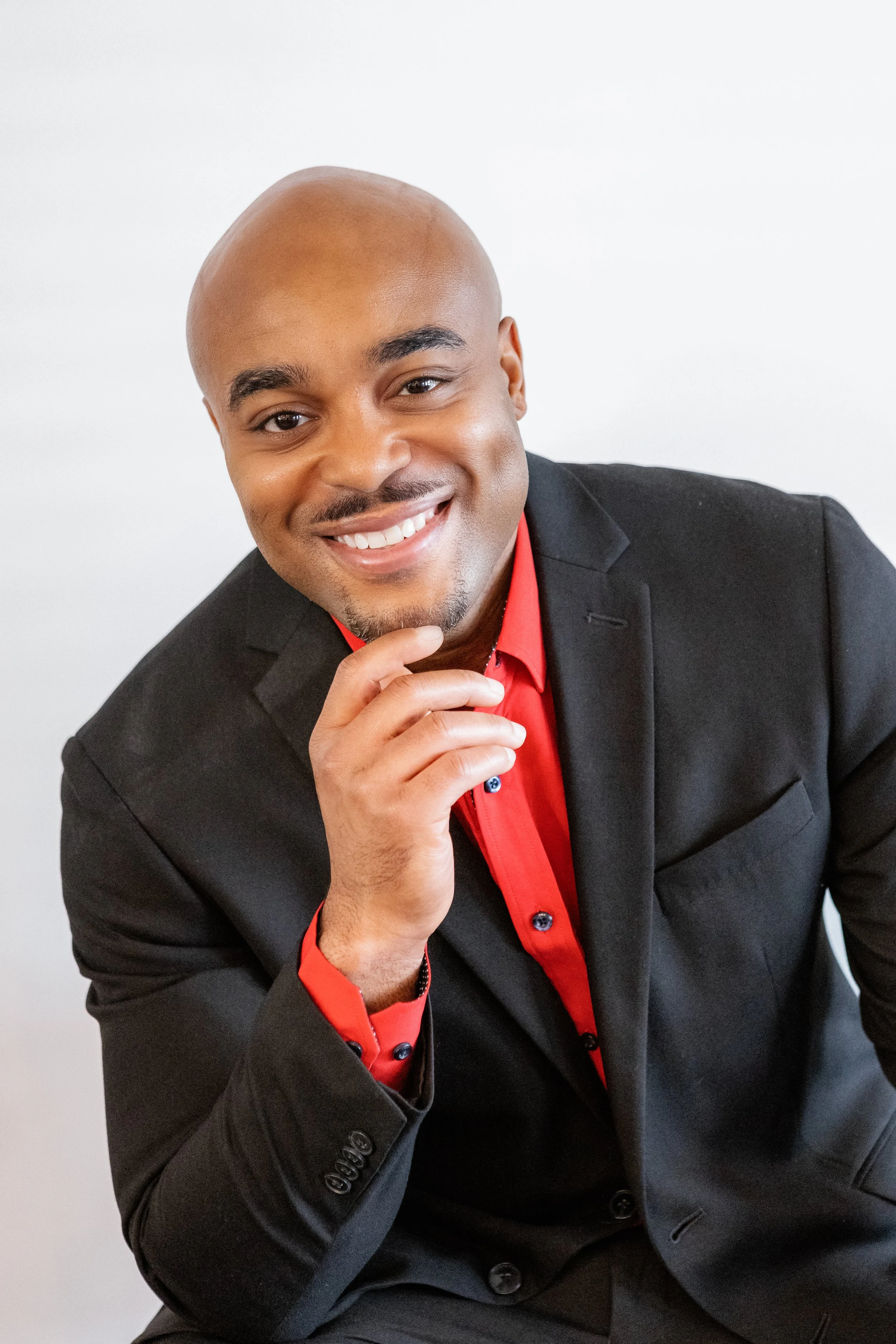 A smiling African American man in a black suit with a red shirt, sitting with his hand touching his chin, against a plain white background.
