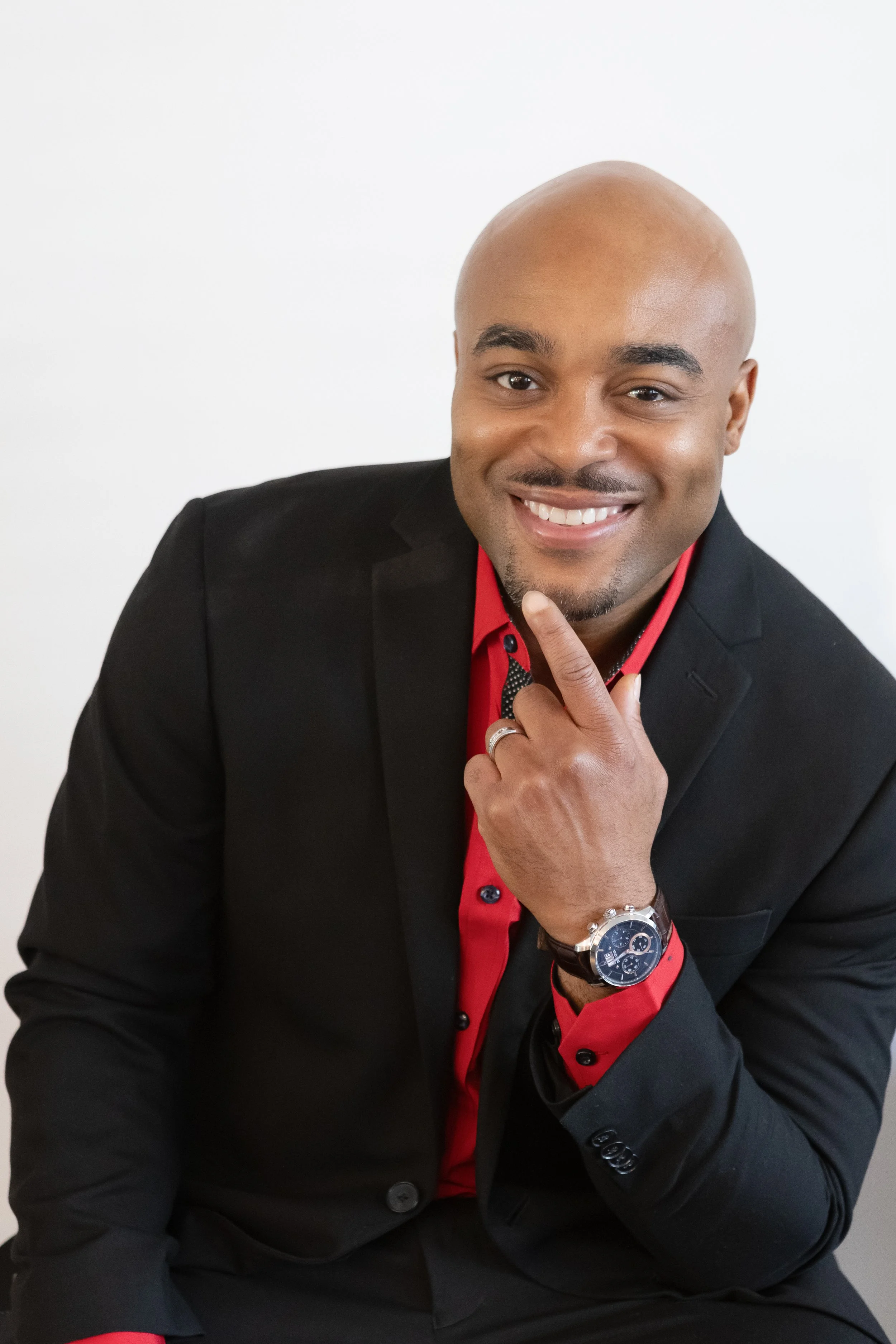Portrait of a bald man in a black suit with a red shirt, smiling and touching his chin, wearing a watch and ring, against a plain white background.