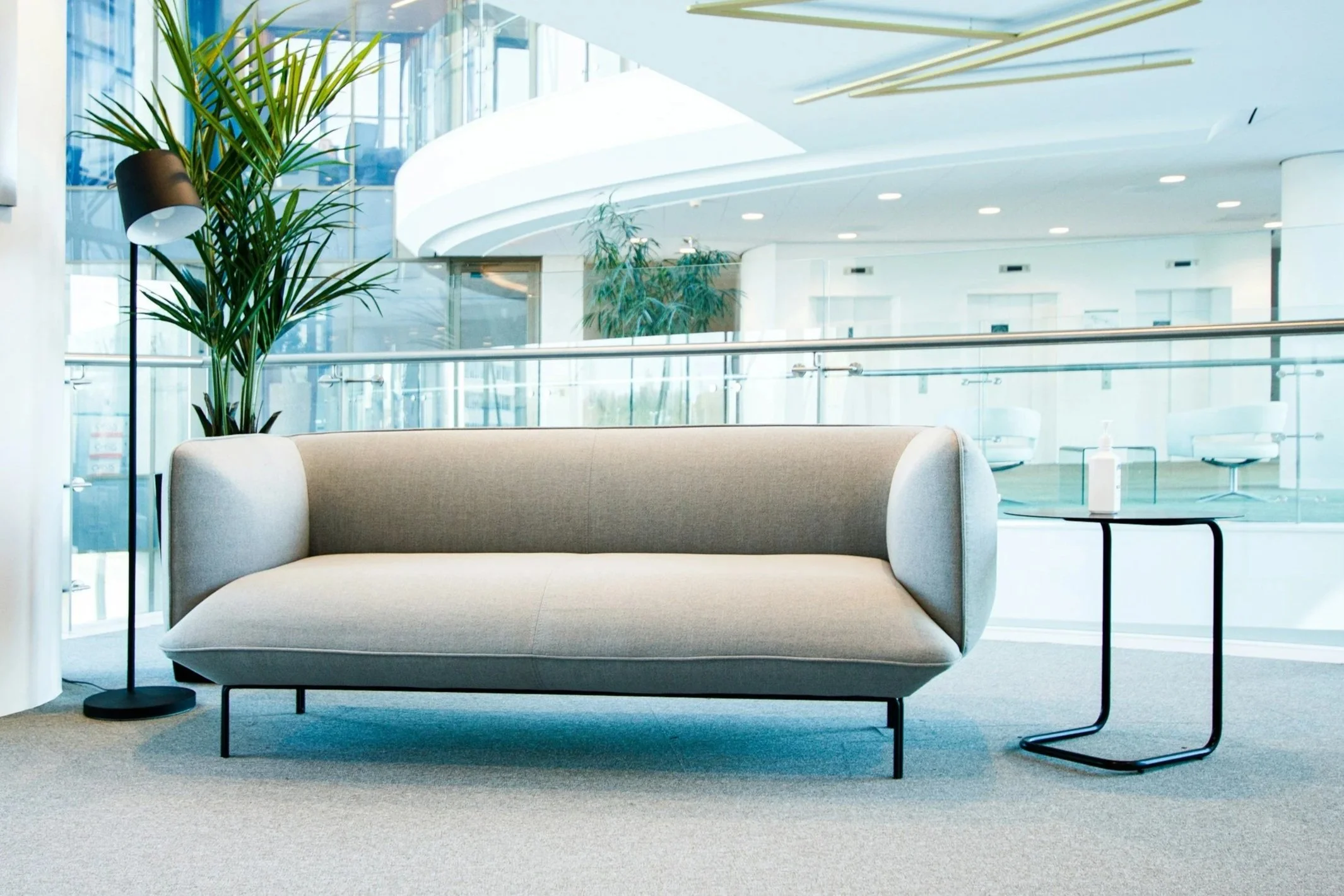 Modern office waiting area with beige sofa, side table with hand sanitizer, tall potted plant, and floor lamp in the background.