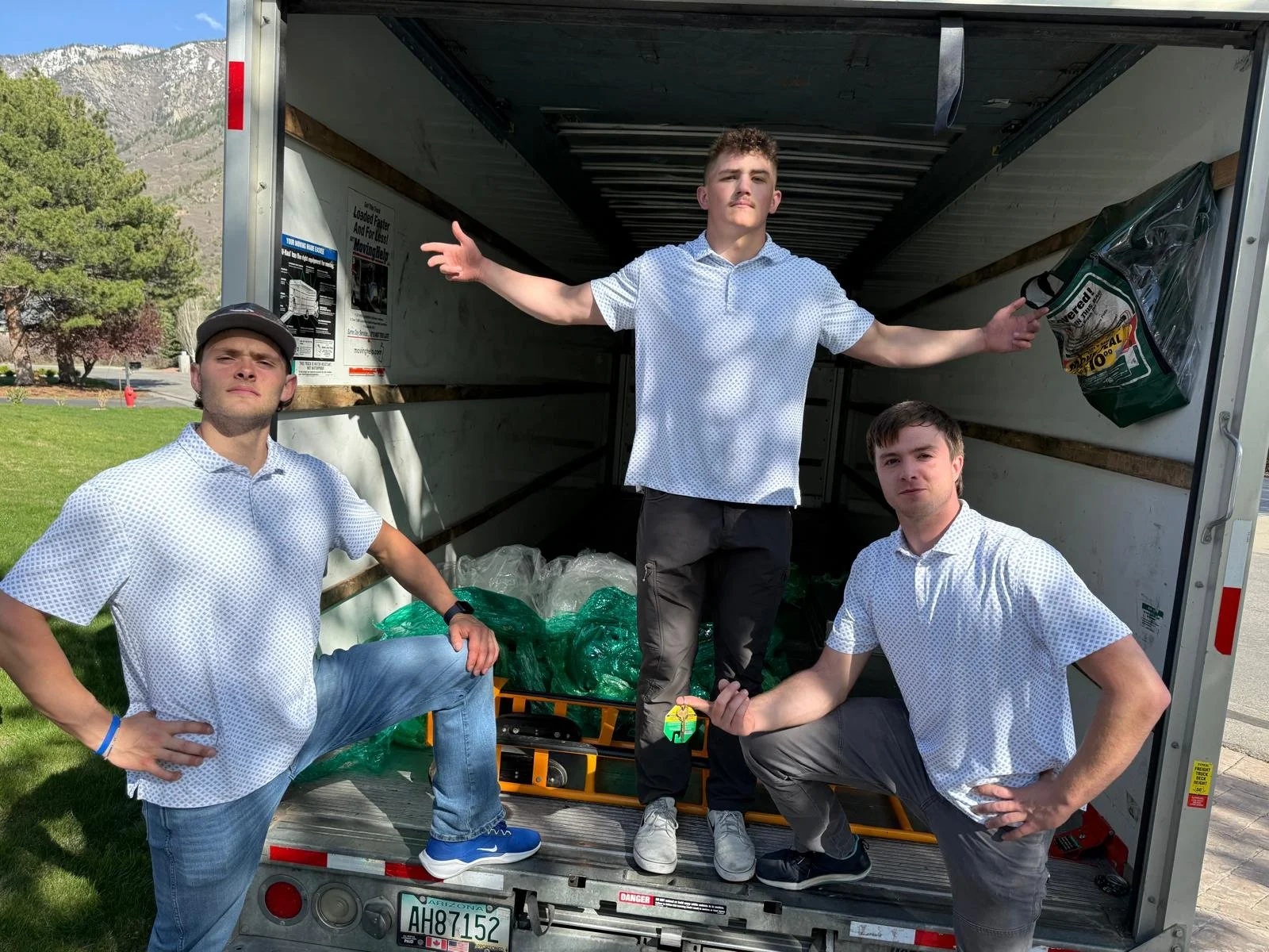 Three young men in white polo shirts and jeans perform playful poses inside an open moving truck filled with green plastic bags, with mountains and trees in the background.