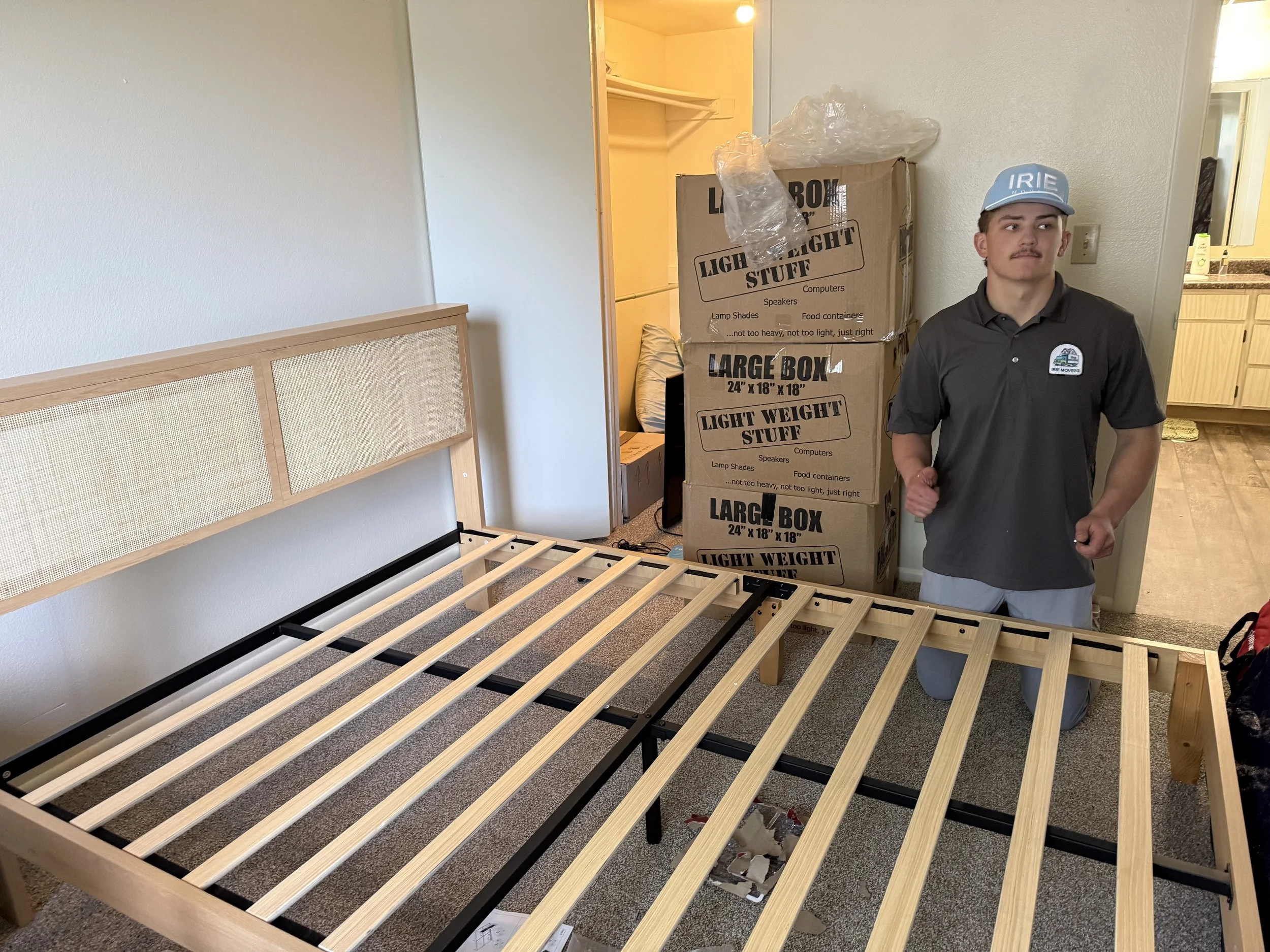Room with a partially assembled bed frame and a person kneeling beside it. The person is wearing a gray polo shirt, light gray shorts, and a blue cap that says 'IRIE.' Two large boxes labeled 'Large Box' and 'Light Weight Stuff' are stacked behind them.