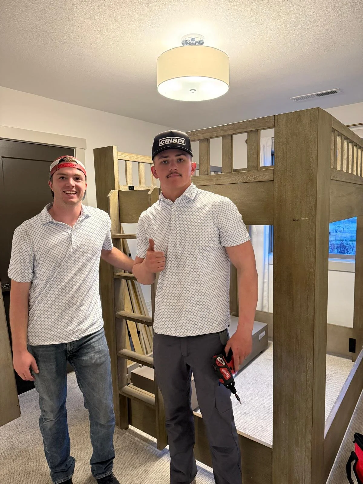Two young men standing inside a room with a wooden bunk bed. One man is smiling and the other is holding a power drill, giving a thumbs-up. The room has a ceiling light, a window, and carpeted floors.