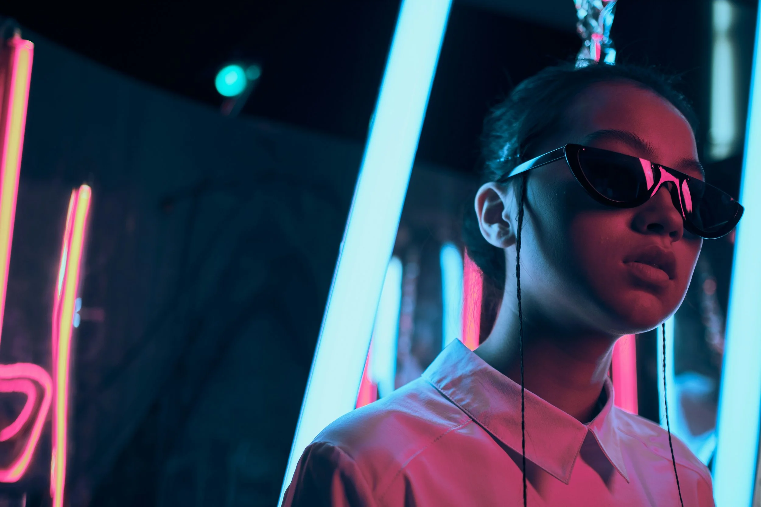 A woman wearing sunglasses and a collared shirt, surrounded by neon lights in a dark setting.