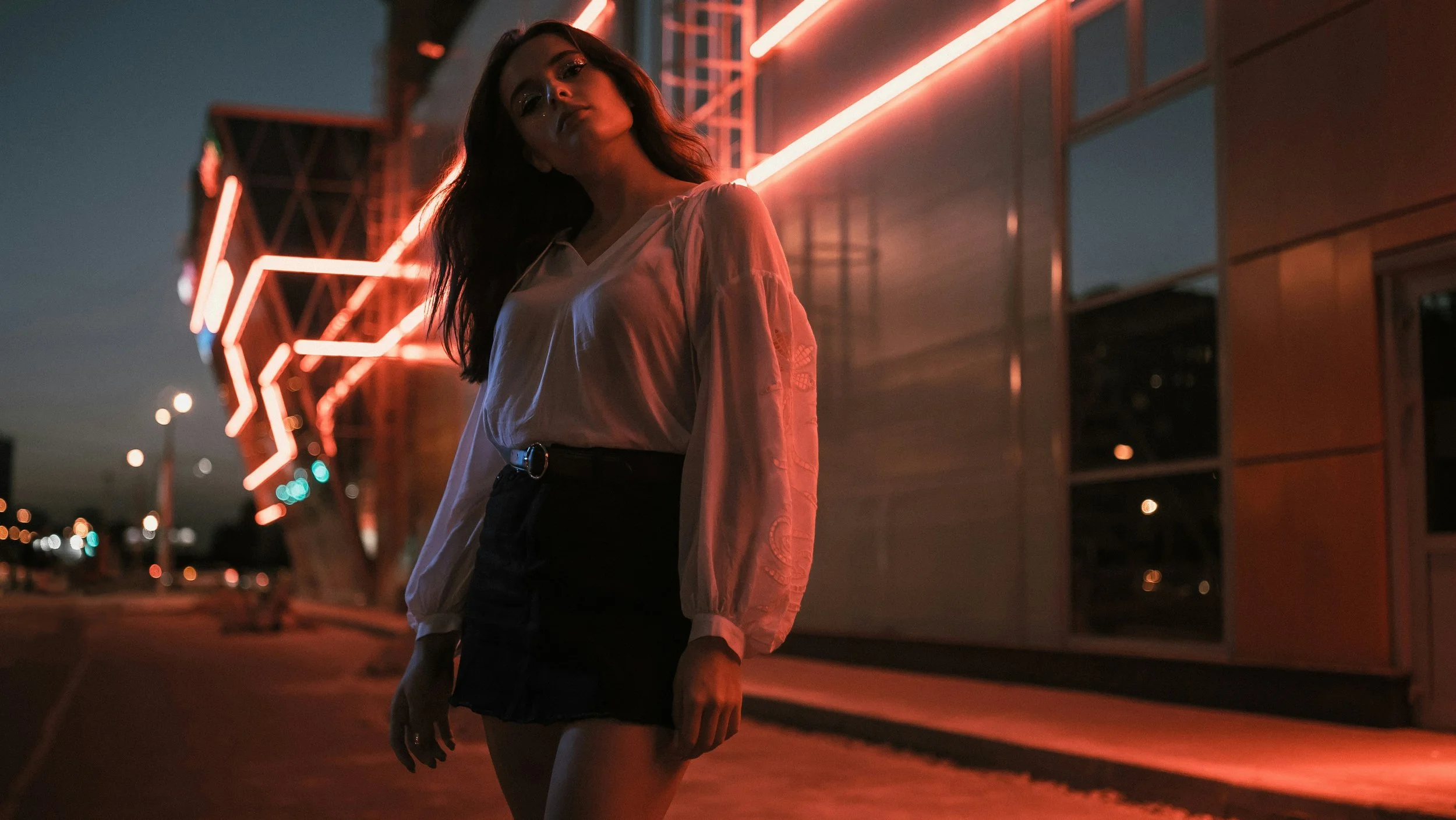 A woman standing outdoors at night in front of a modern building with red neon lights. She wears a white blouse and a black skirt, with her dark hair loose. The scene is dark with city lights in the background.