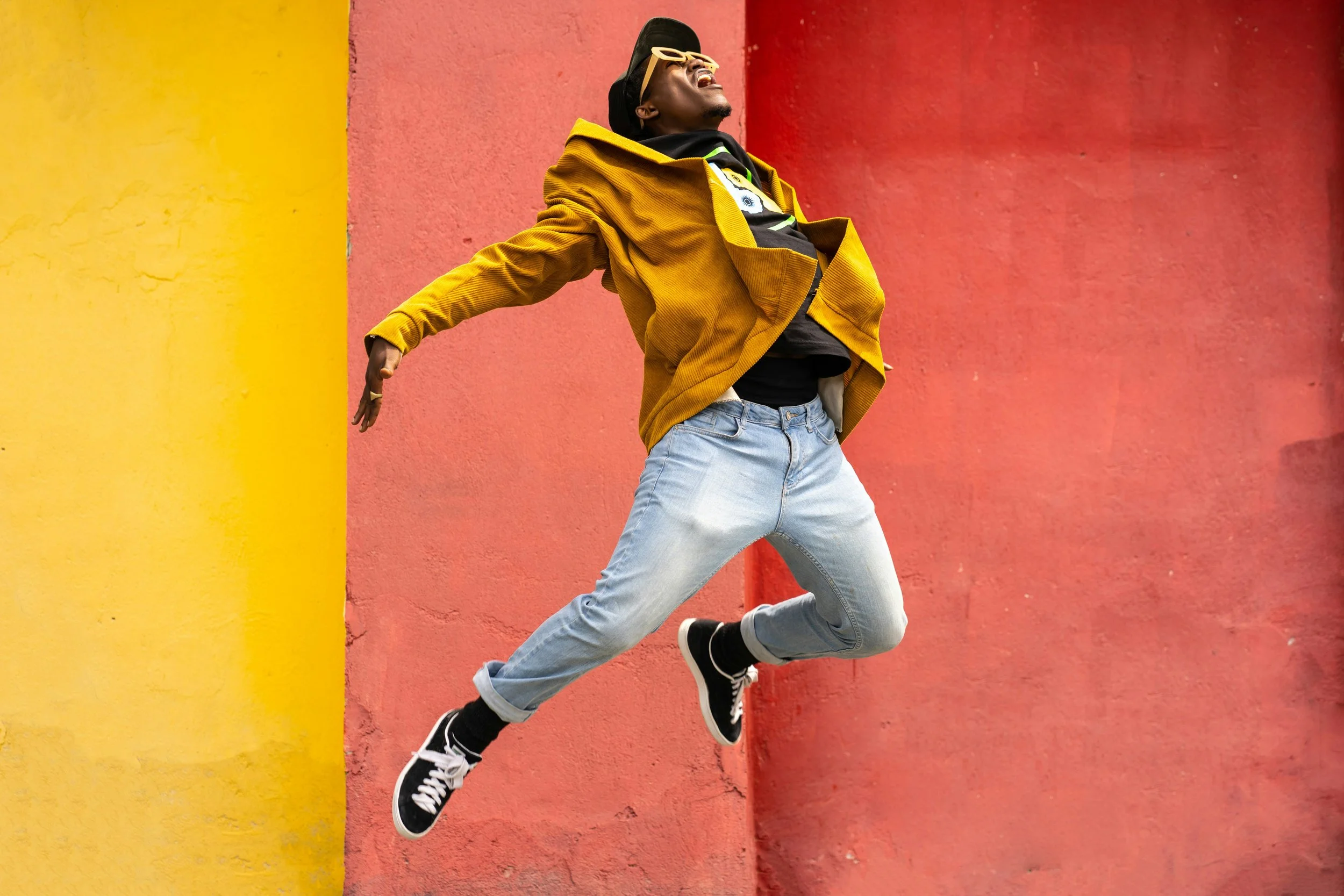 Person mid-air jumping in front of a yellow and red wall, wearing a yellow jacket, black hoodie, ripped jeans, and black sneakers.