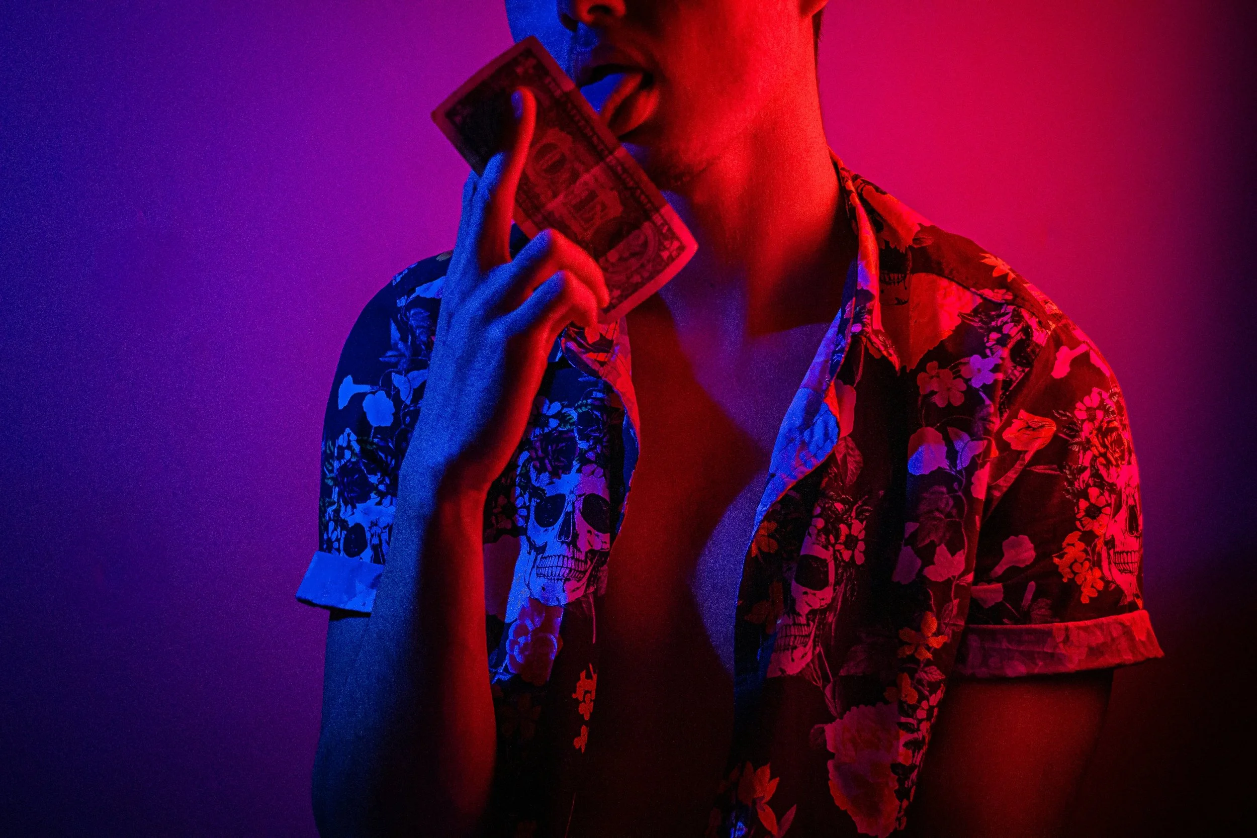 Person holding a $100 bill near their face with their tongue out, wearing a floral patterned shirt under colorful lighting.