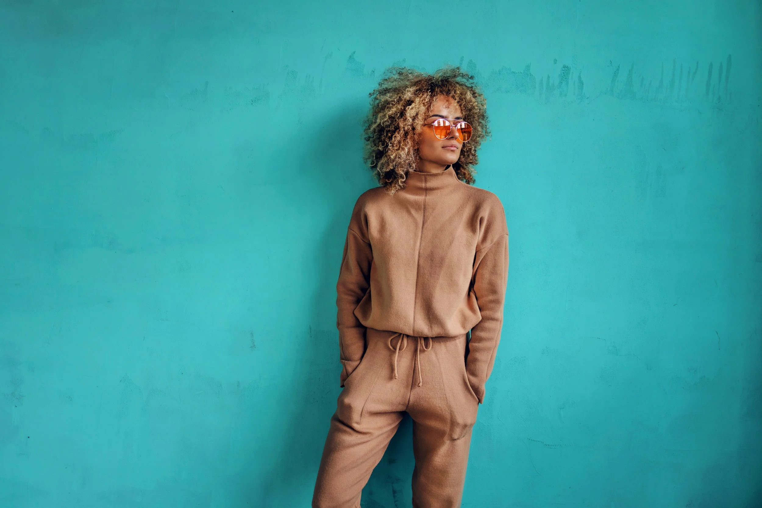 Woman in tan tracksuit with hands in pockets, wearing orange sunglasses, standing against a turquoise wall.