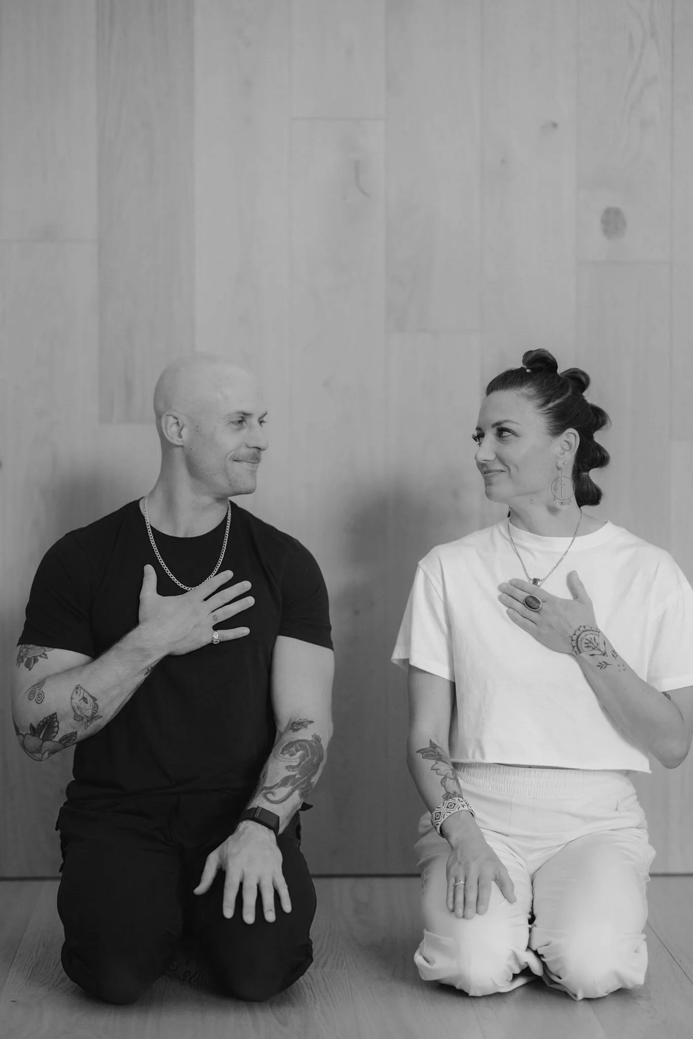 A man and a woman kneeling on the floor, facing each other with their right hands over their chests, smiling and looking into each other's eyes.